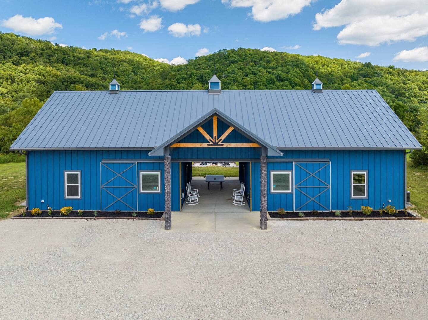 A bright blue barn-style building with a metal roof and open center breezeway, set against a green, wooded hillside.