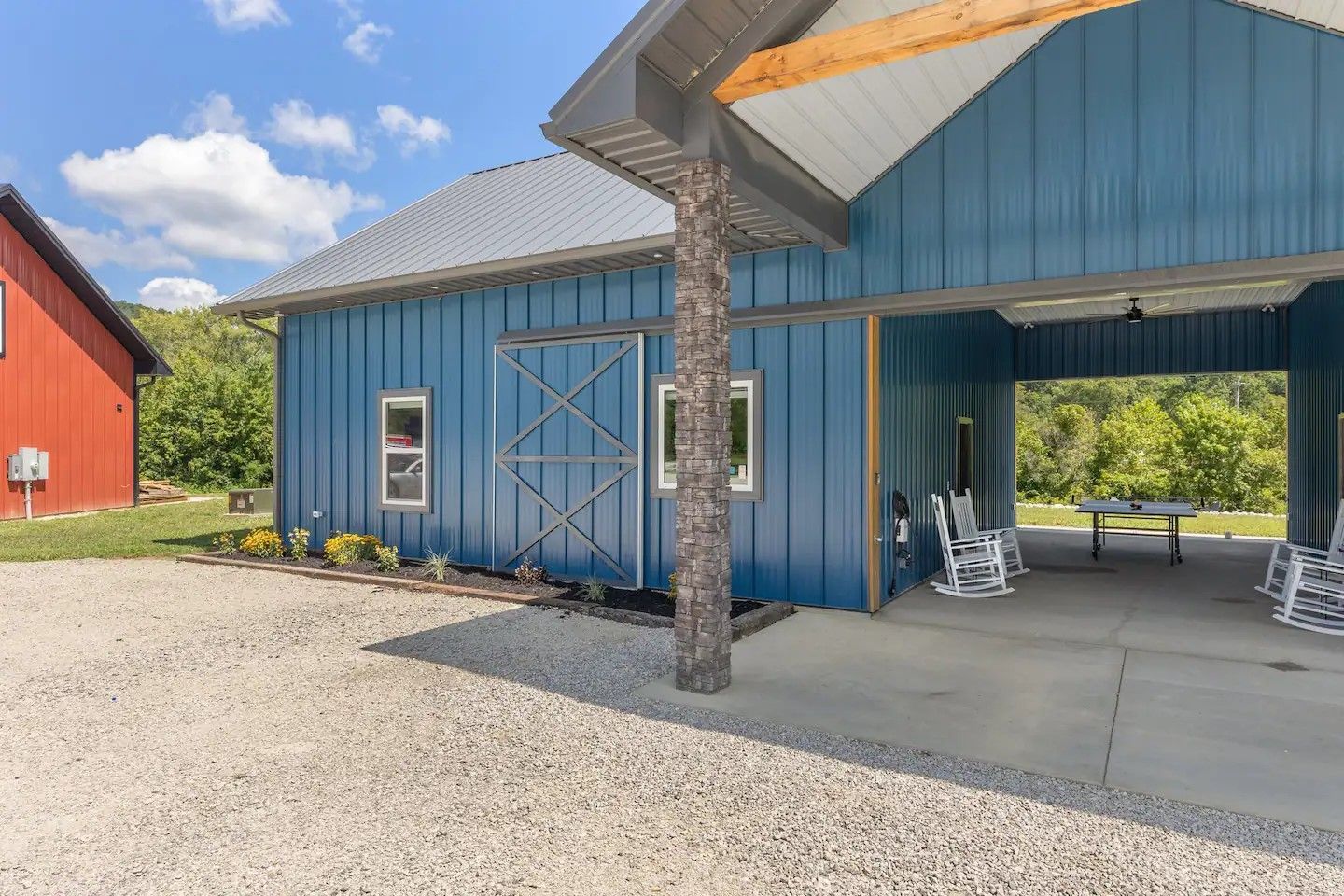 A blue barn-style building with a gravel driveway, covered porch with chairs, and a red building in the background.