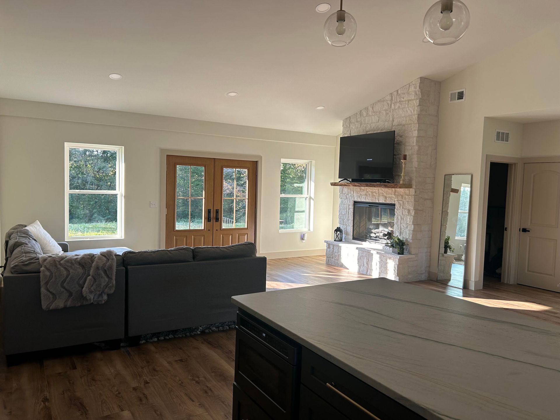 A spacious open-plan living room with a grey sectional, fireplace, white brick wall, and dark kitchen island in foreground.