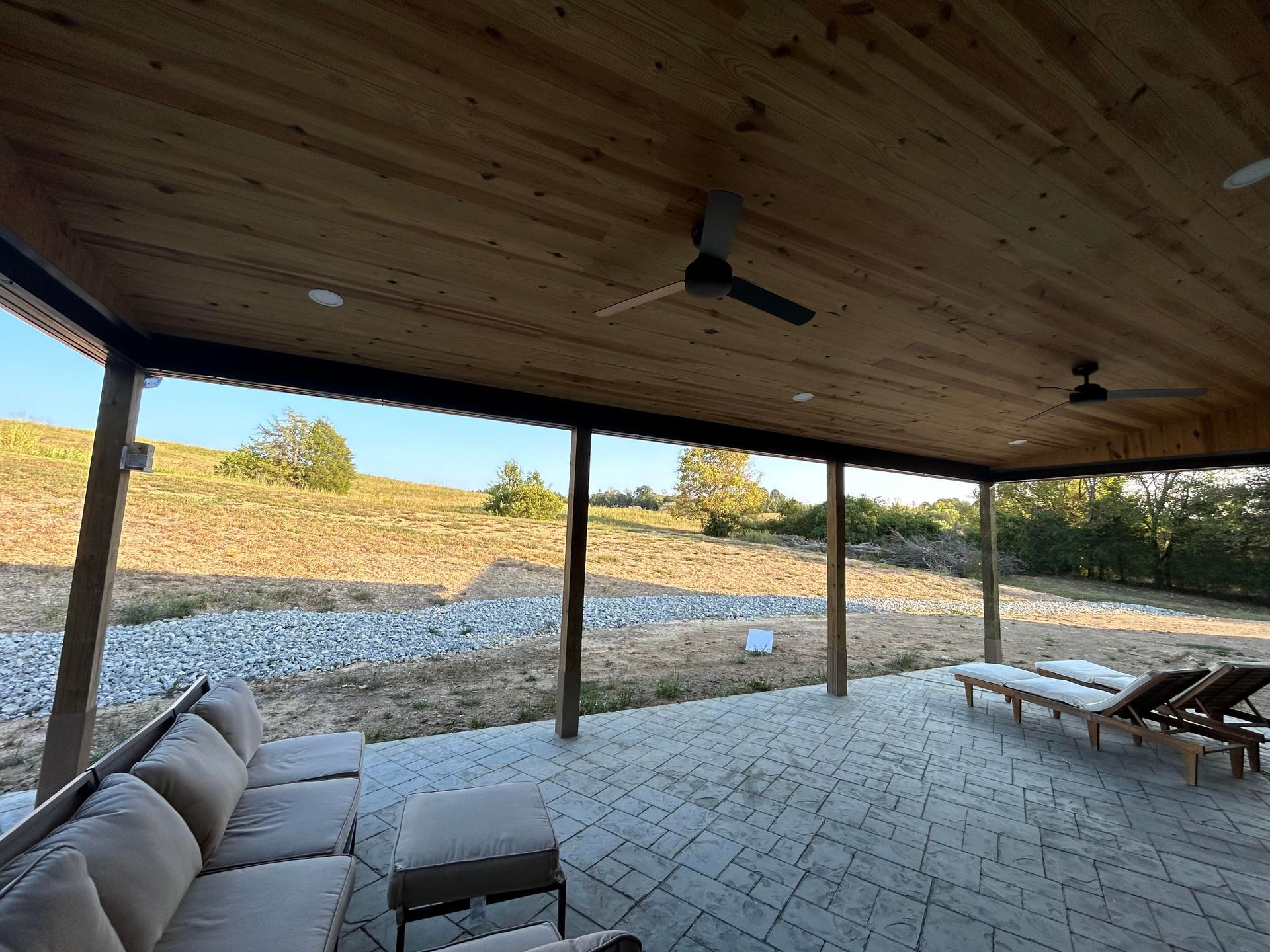 A covered outdoor stone patio with comfortable seating, ceiling fans, and a view of a grassy hill.