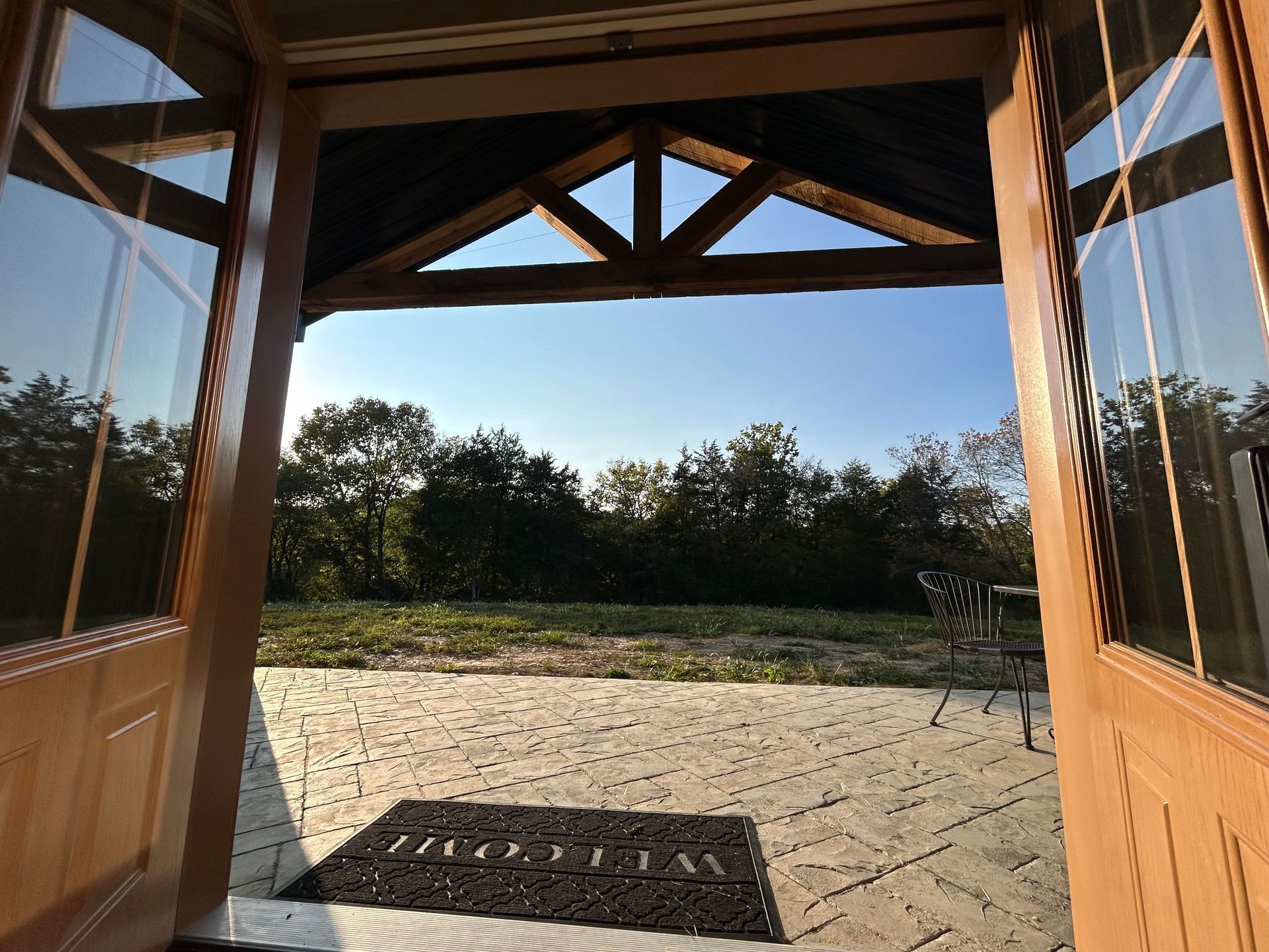 A view through open wooden doors onto a stone patio with a welcome mat, looking out toward trees under a blue sky.