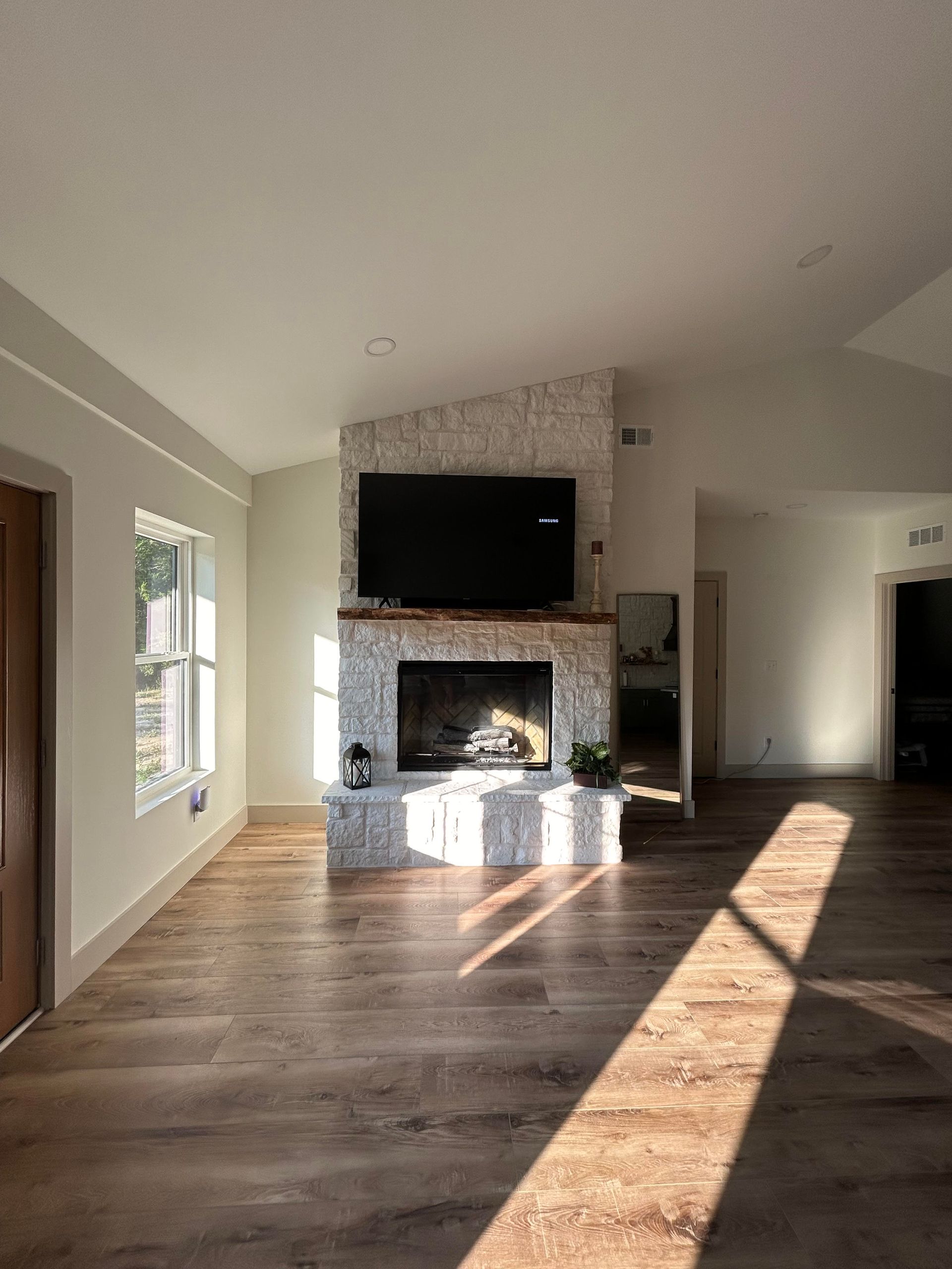 A living room features a white stone fireplace with a wall-mounted TV and a natural wood mantel on light wood floors.