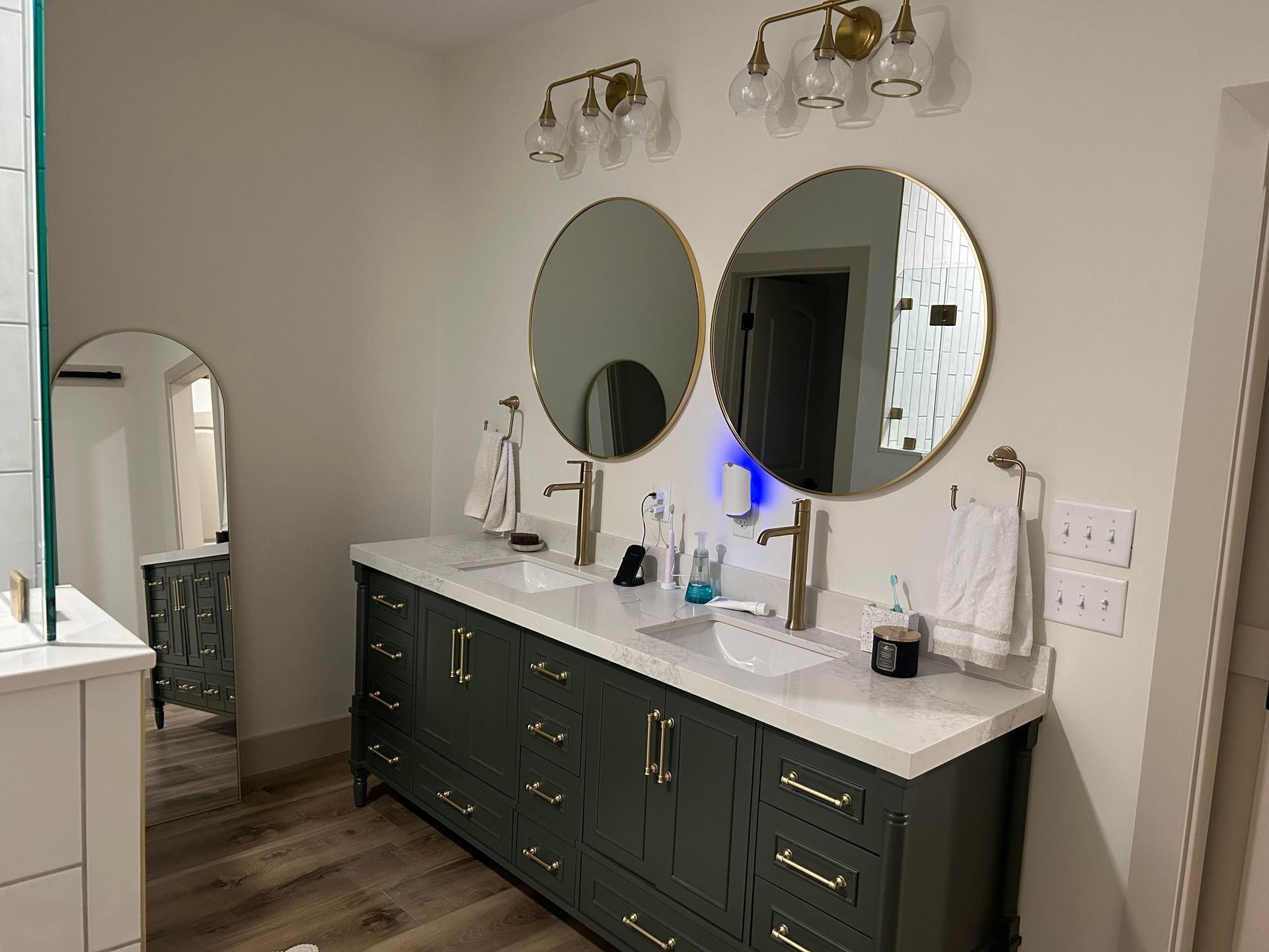 A bathroom with a dark green double vanity, gold hardware, two round mirrors, and brass light fixtures.