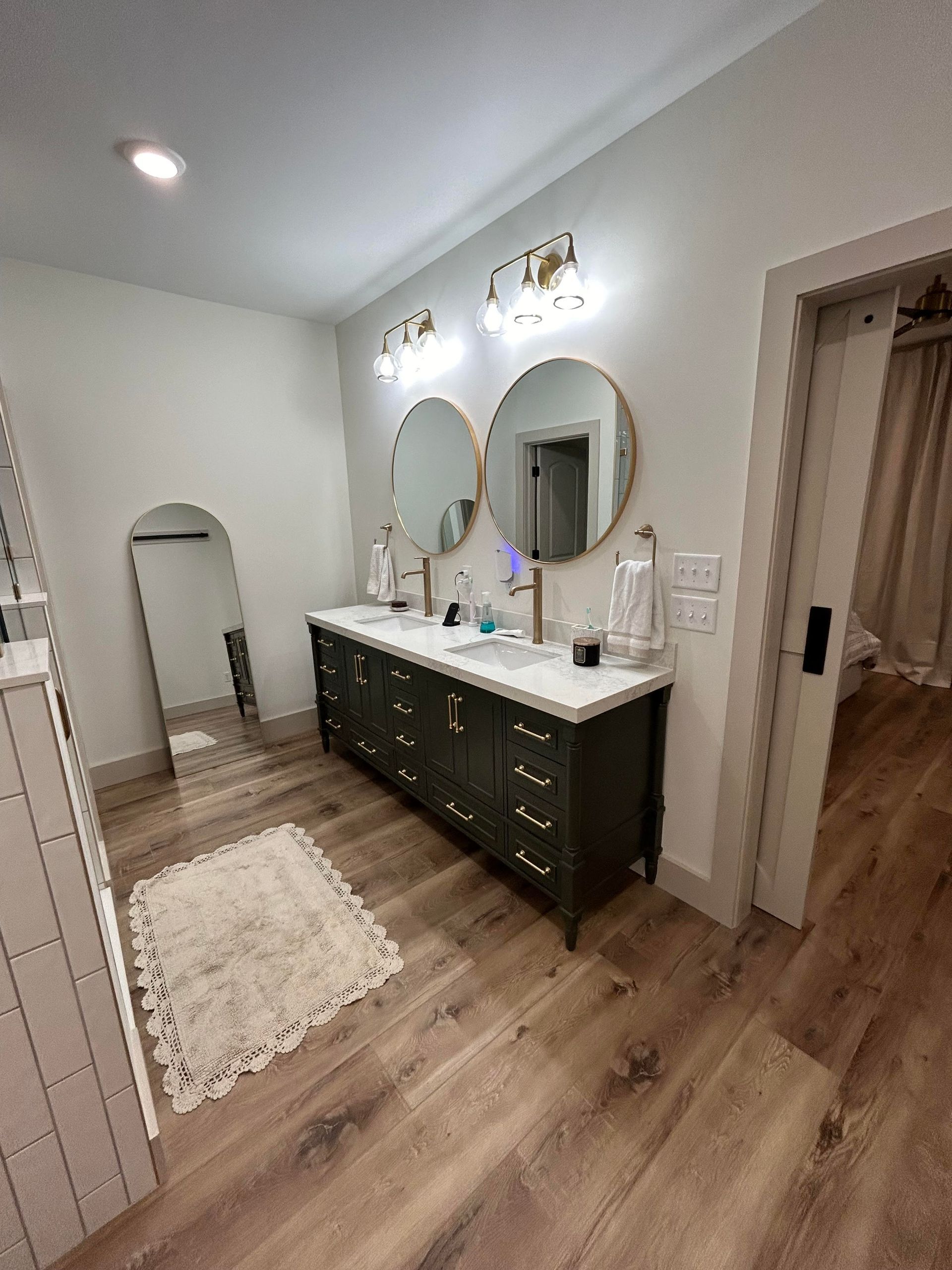 A bathroom with a dark double vanity, two round mirrors, gold light fixtures, an arched floor mirror, and wood floors.