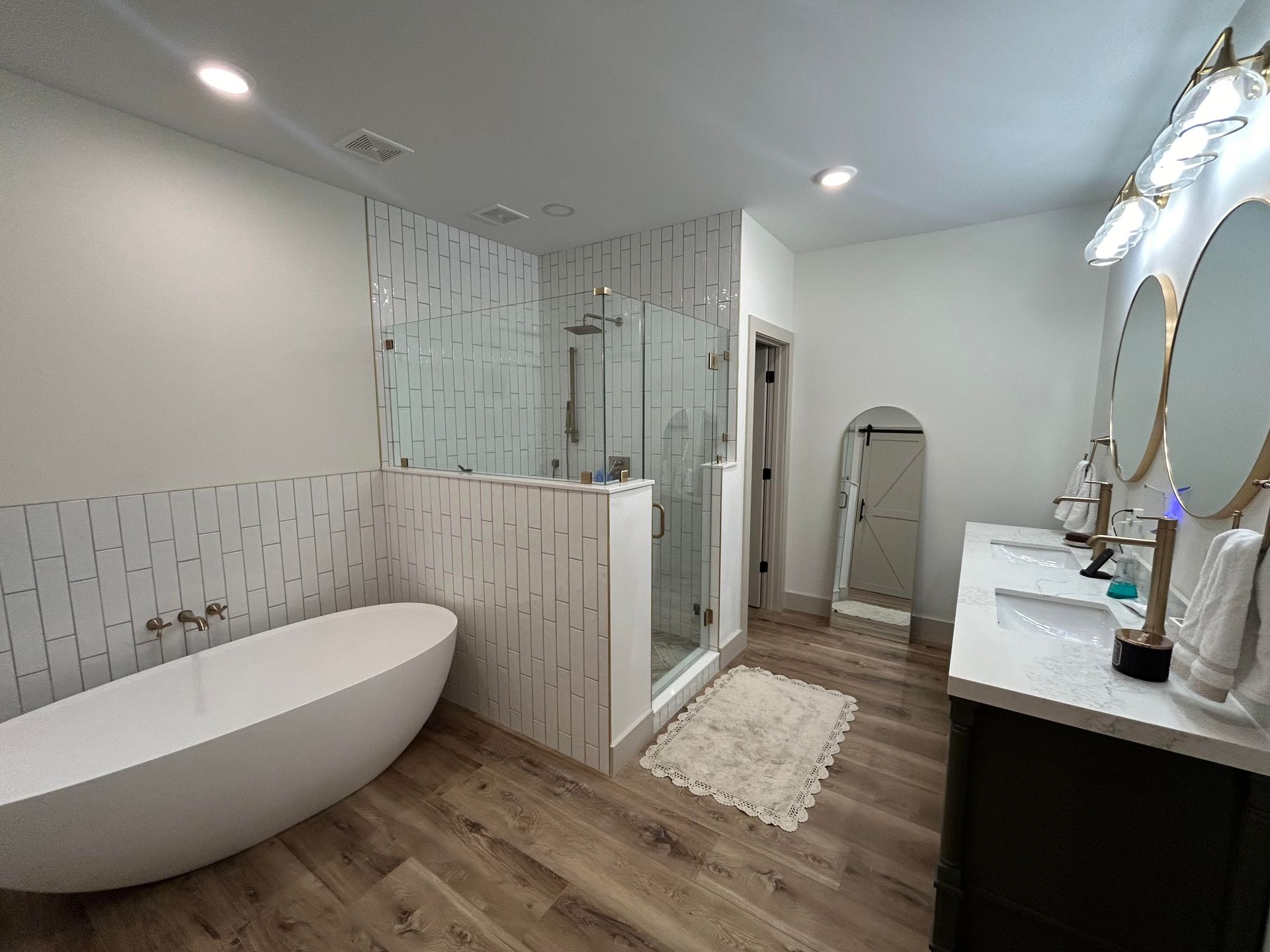 A modern bathroom featuring a white soaking tub, a tiled walk-in glass shower, double vanity, and wood-look flooring.