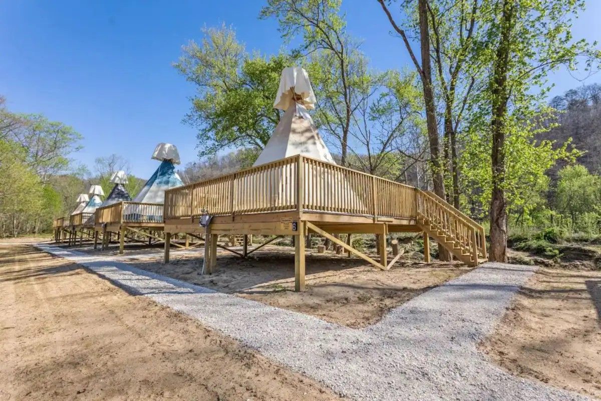 A row of teepee-style tents on elevated wooden decks set in a sunny, wooded outdoor area with a gravel path.