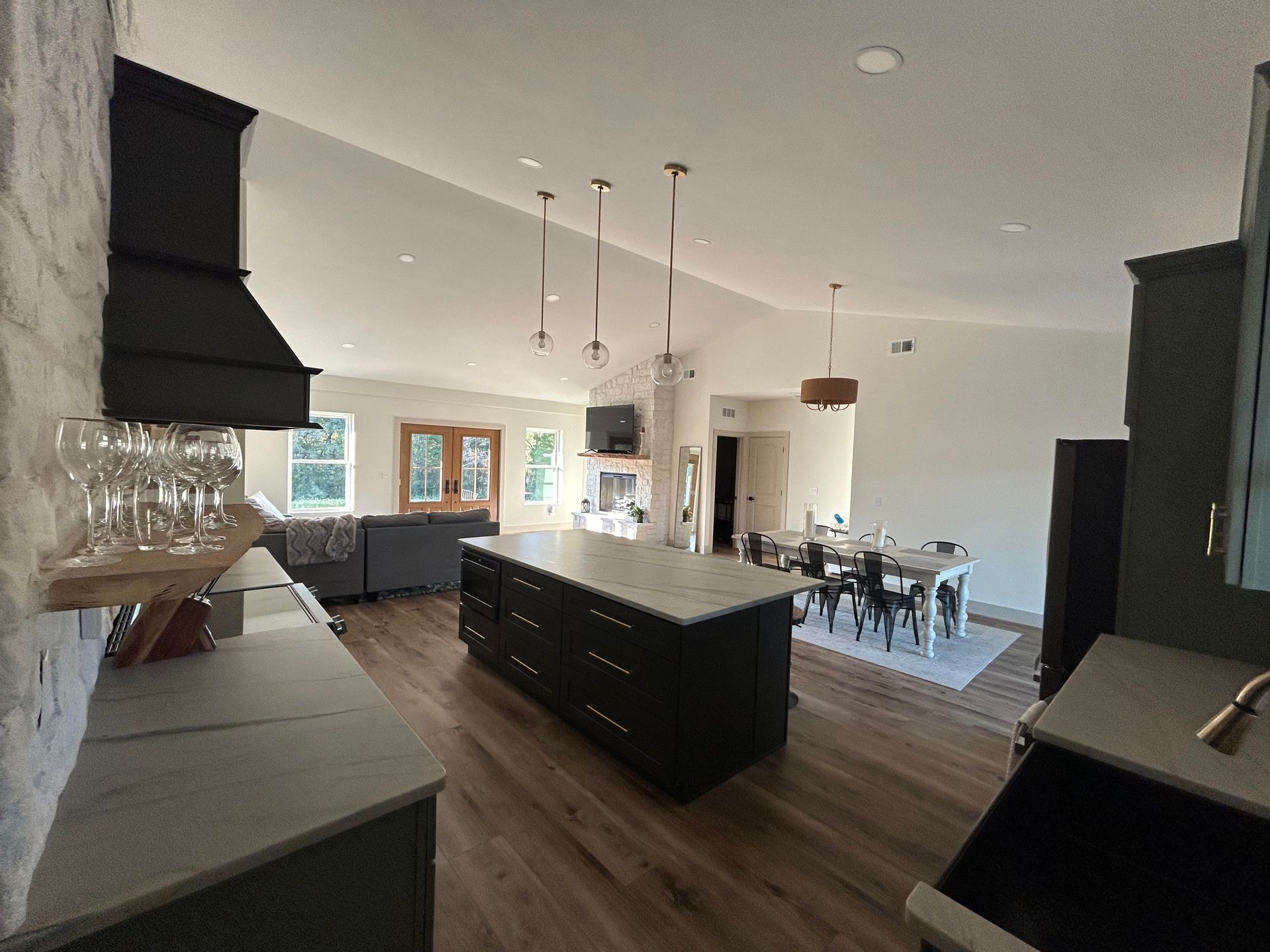 A modern open-plan kitchen and dining area with a large black island, stone walls, wood floors, and a bright living space.