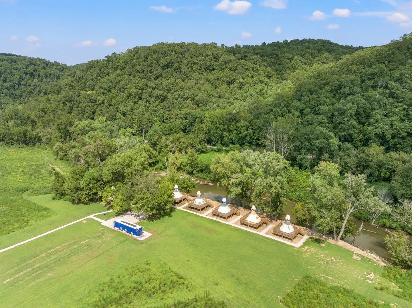 An aerial view of five white stupas set in a grassy field next to a tree-lined stream, with a small blue building nearby.