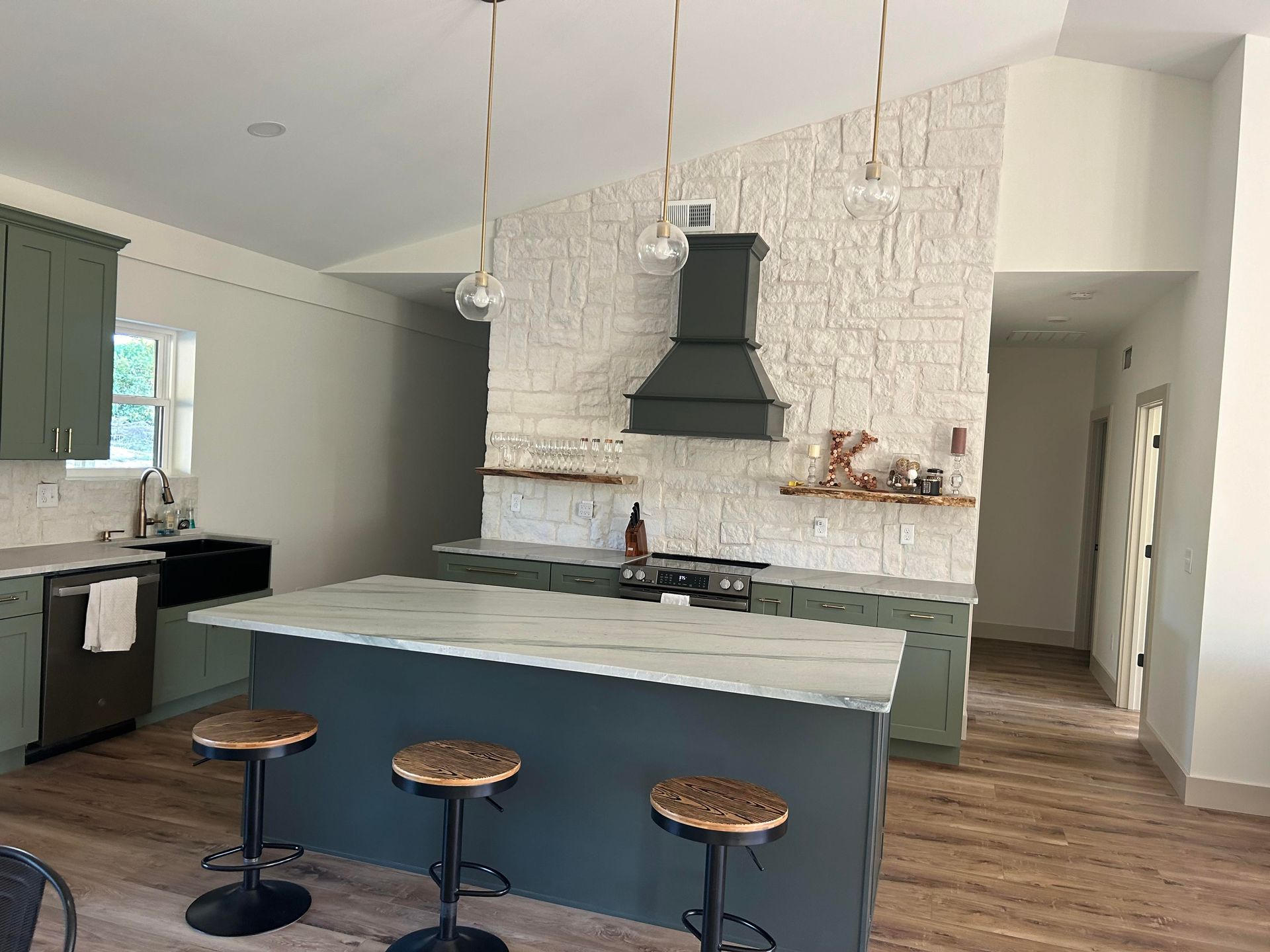 A kitchen island with three stools sits in front of a white stone wall featuring a dark range hood and wooden shelving.