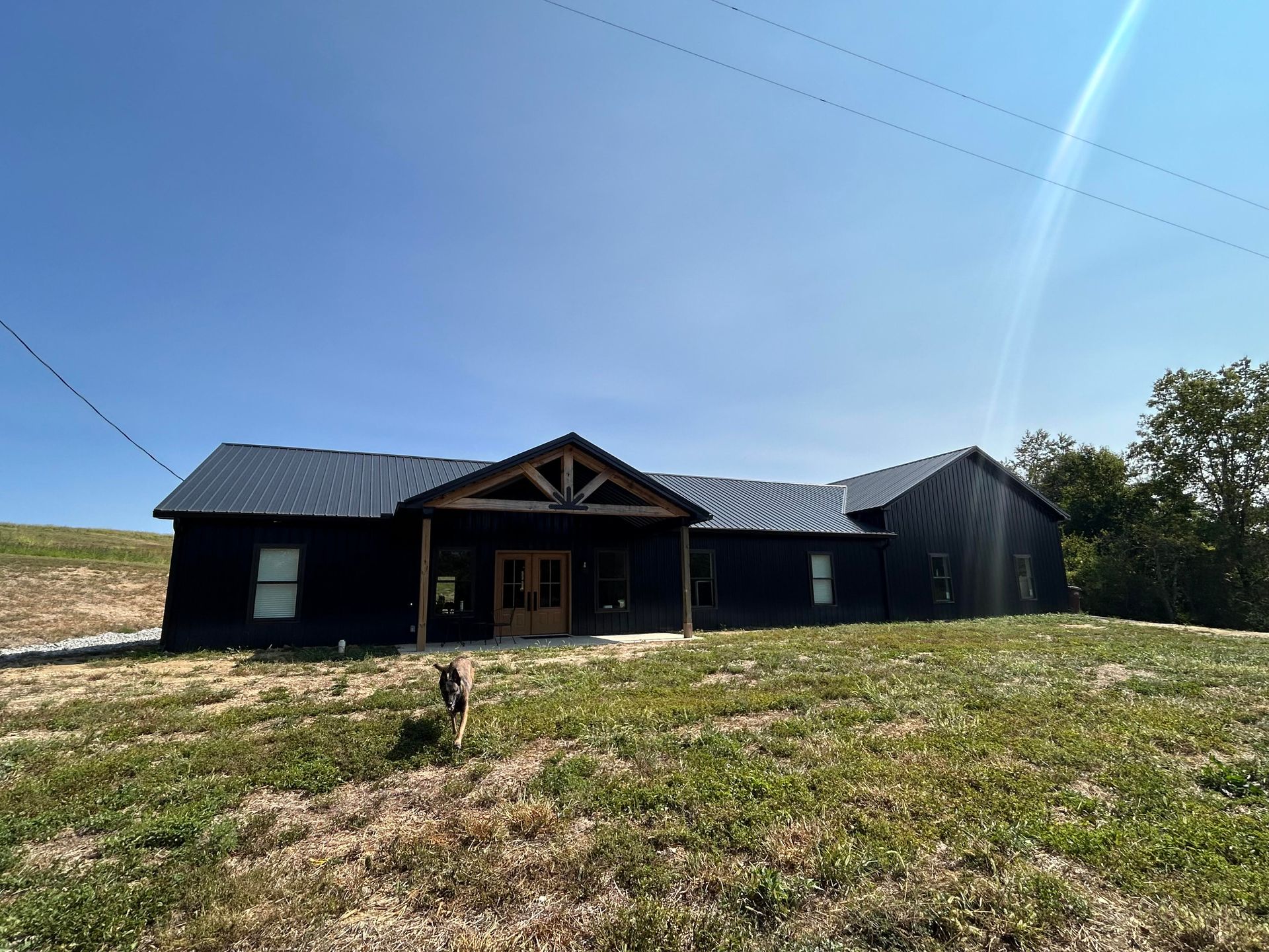 A modern, dark-sided single-story house with a wooden entryway sits on a grassy hill under a bright blue sky.