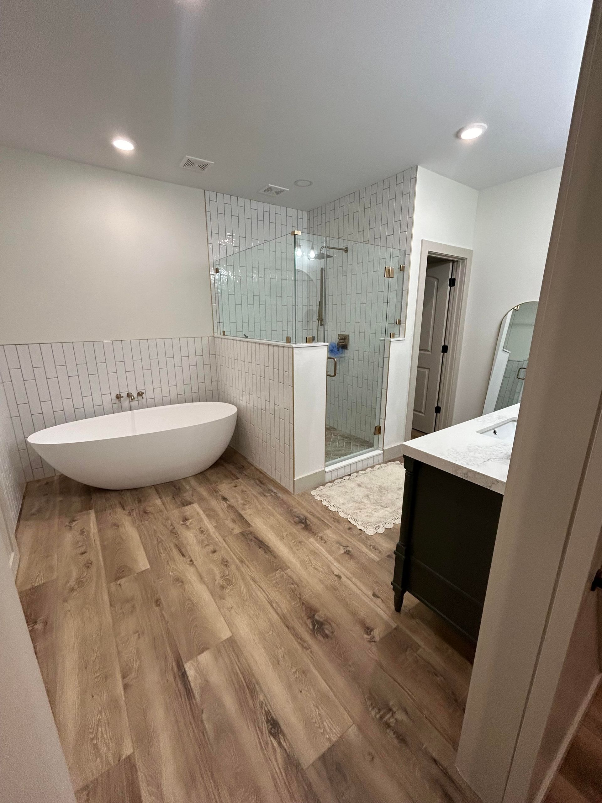Modern bathroom with wood-look flooring, a white freestanding tub, a glass-enclosed shower, and a dark vanity.