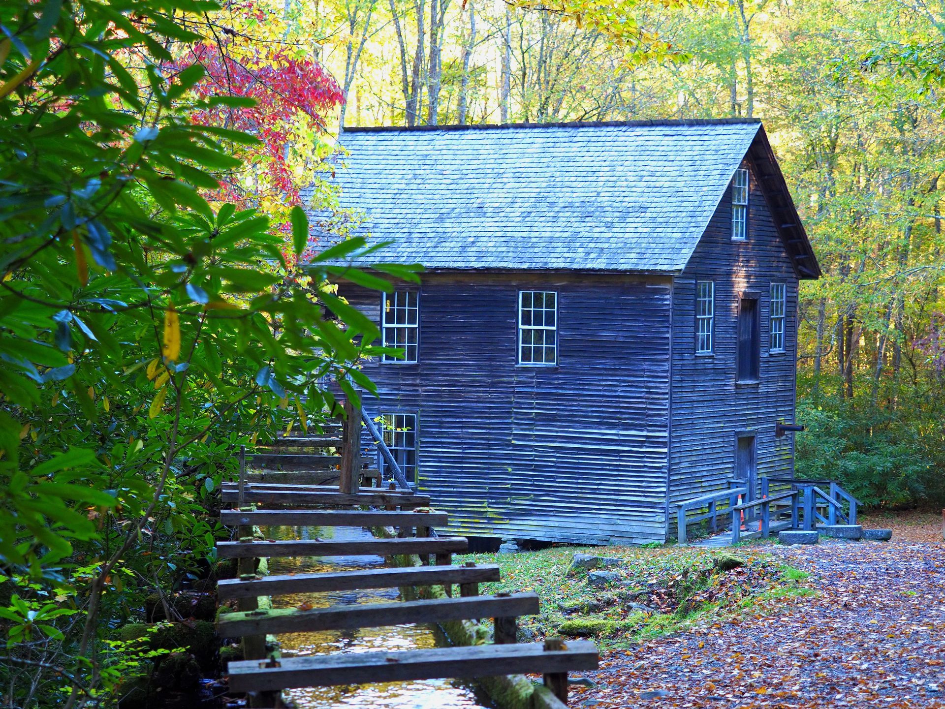 Old wooden mill with water flowing through, surrounded by fall foliage.