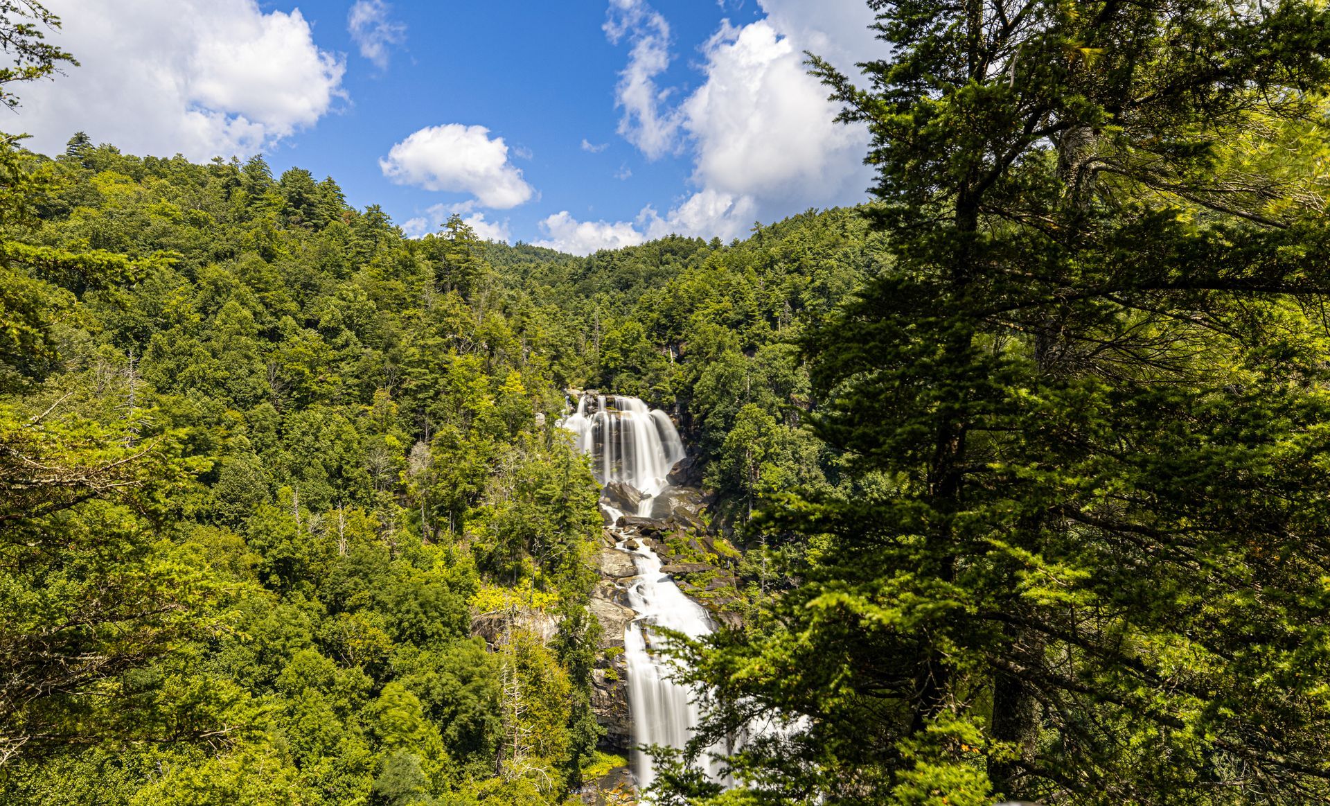 Waterfall cascading down a rocky cliff, surrounded by lush green trees, under a blue sky with clouds.