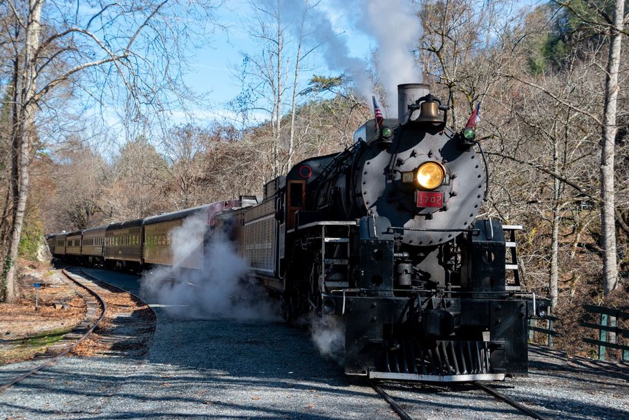 Steam train traveling on tracks through a wooded area, releasing steam.