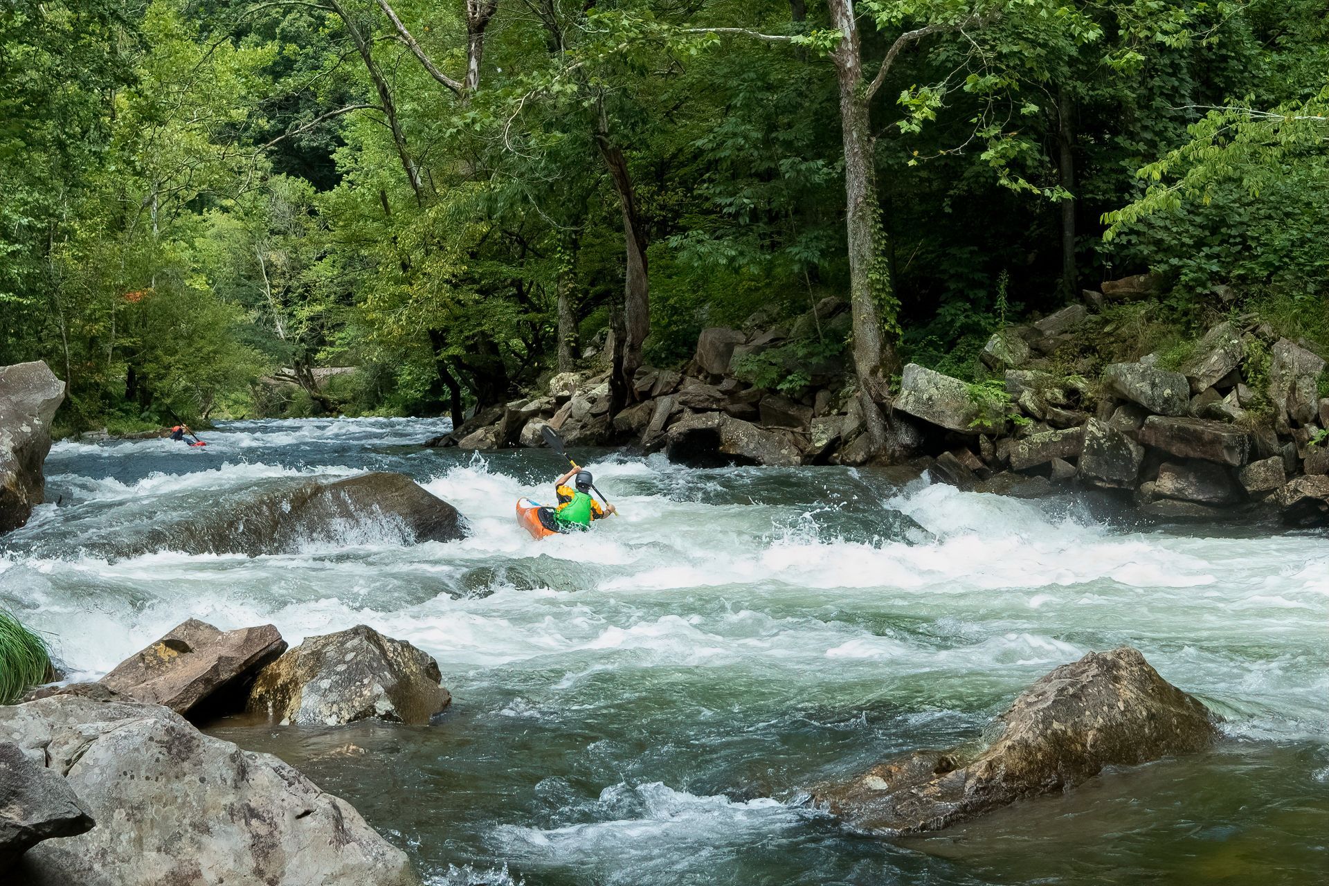 Kayaker navigates white water rapids in a rocky river surrounded by lush green trees.