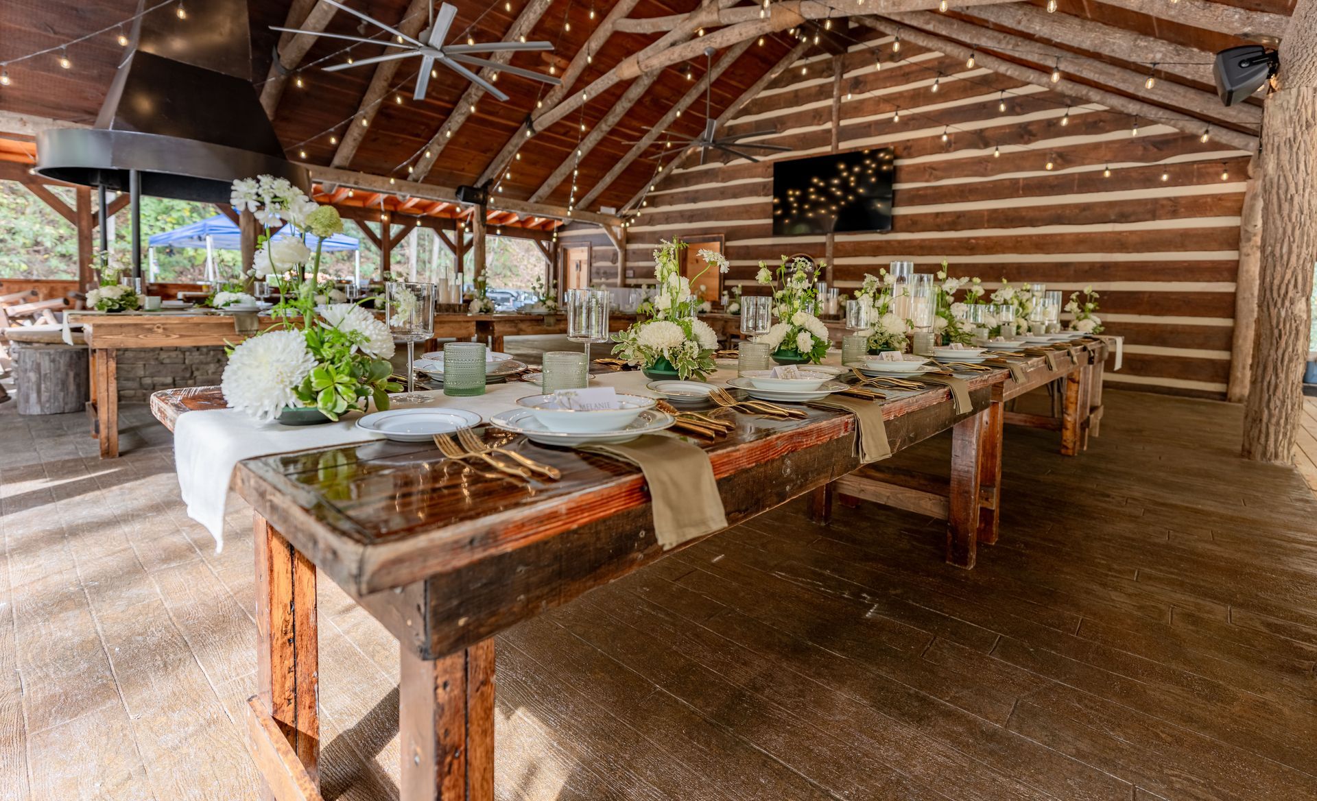 Long wooden table set for a formal gathering, under a log-structured pavilion, decorated with white floral arrangements.