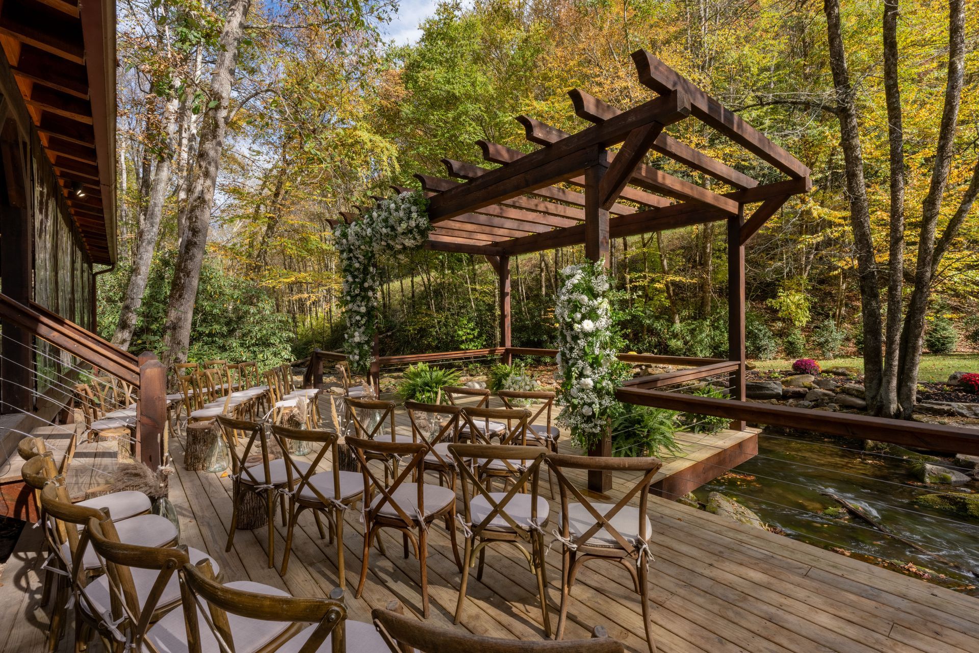 Wooden chairs arranged for an outdoor event next to a pergola with autumn trees in the background.