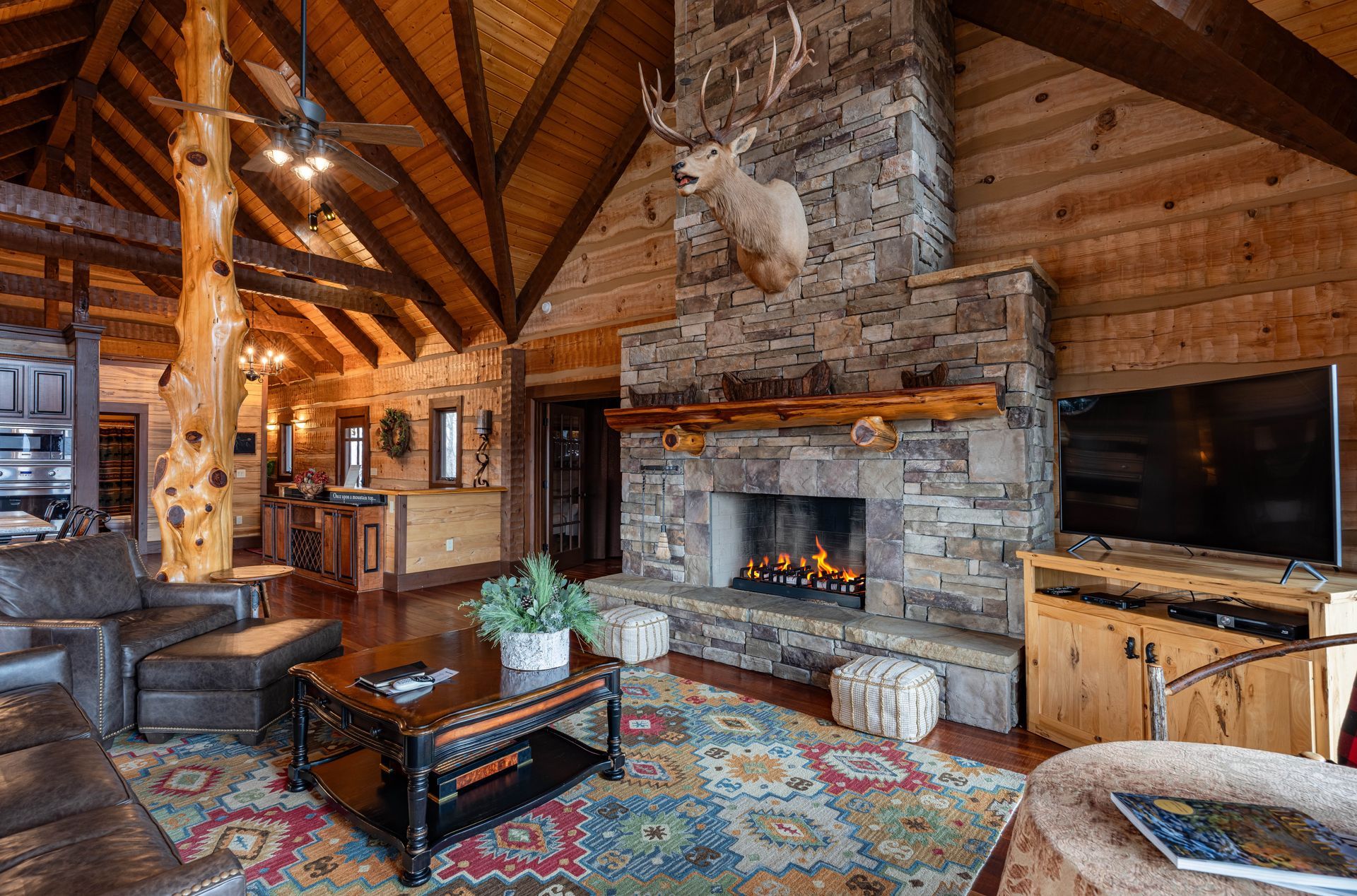 Cozy cabin living room with stone fireplace, leather sofa, wood beams, and a mounted deer head.