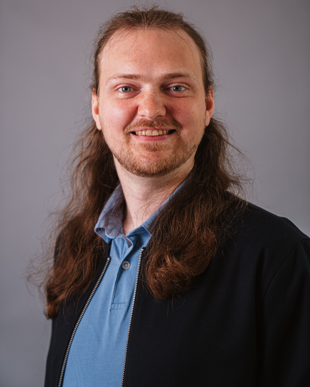 Smiling person with long hair in a blue shirt and dark jacket against a gray background