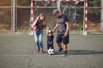 Three people stand on a soccer field by a goal, with a child kicking a soccer ball.