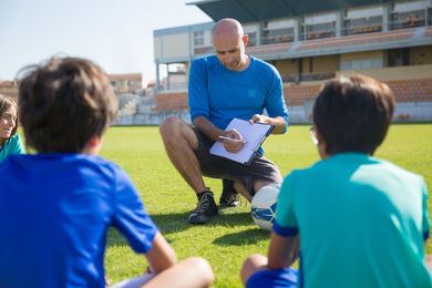 Coach crouches on a field, reading papers to seated children during an outdoor sports discussion