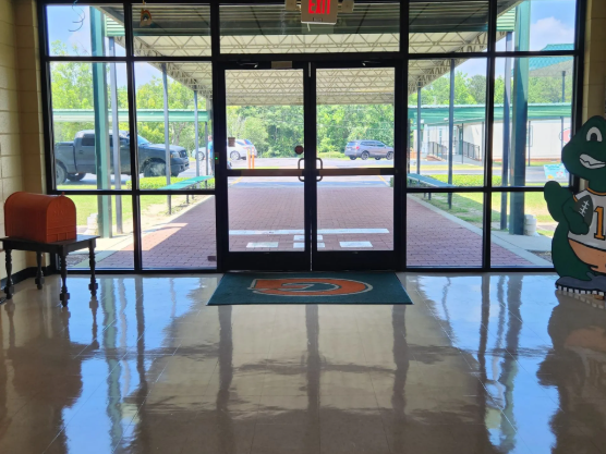 Empty building lobby with glass doors, glossy floor reflections, and a green mascot cutout on the right.