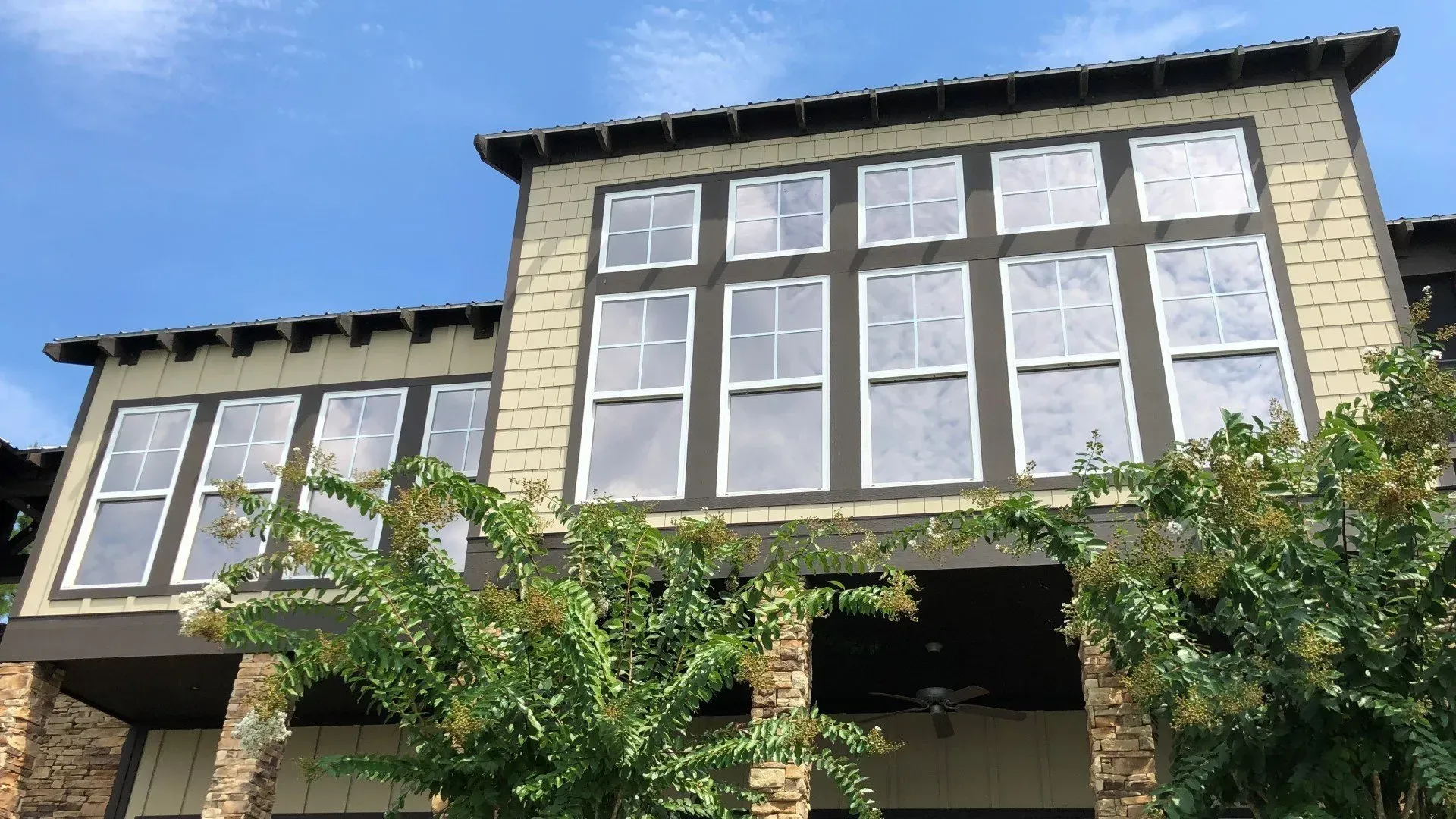 Two-story beige house with large windows and greenery in front, seen from below against a blue sky