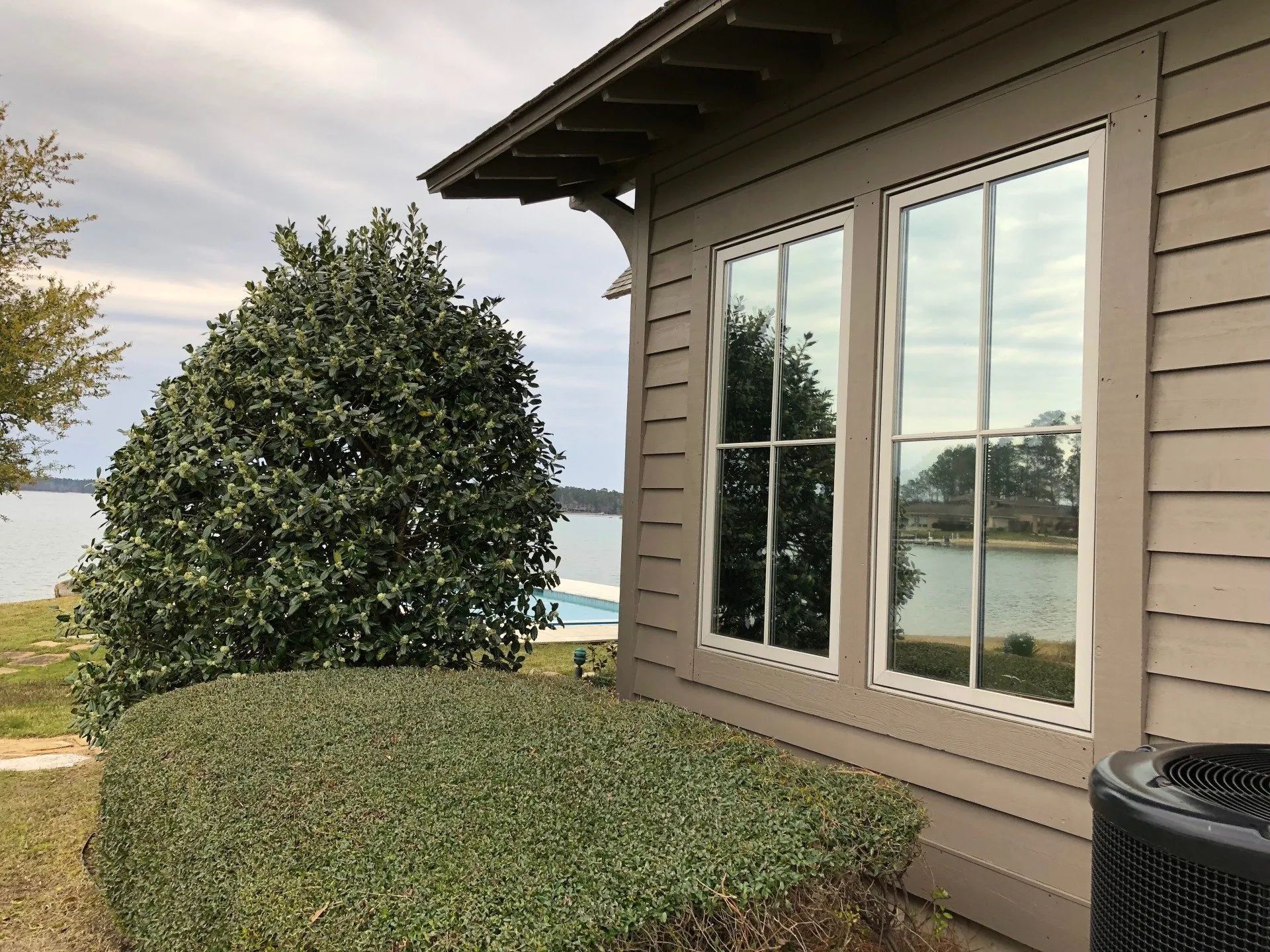 House exterior with beige siding, large windows, trimmed shrub, and a lake view in the background