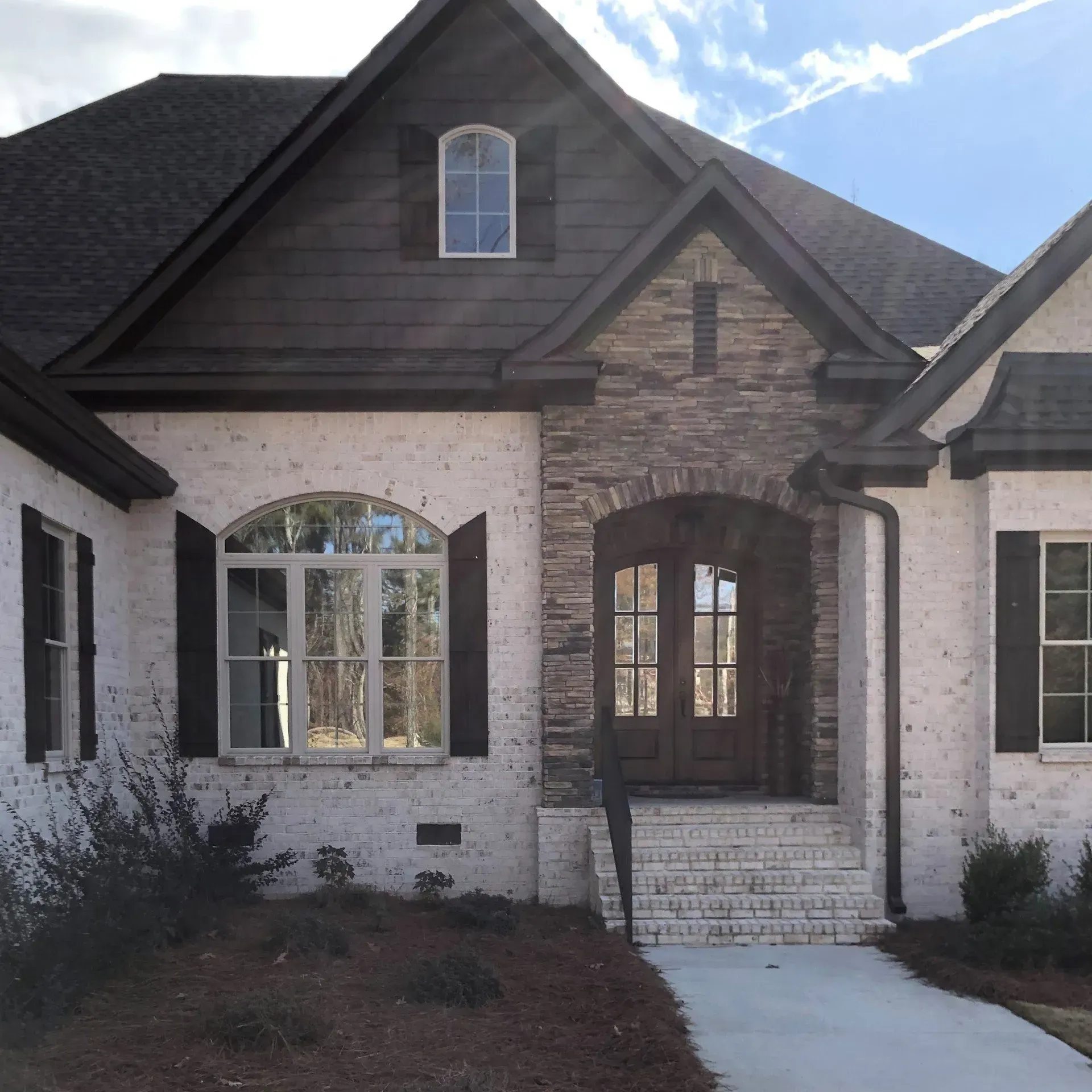 Front view of a white brick house with dark roof and arched wood entryway, lit by sunlight