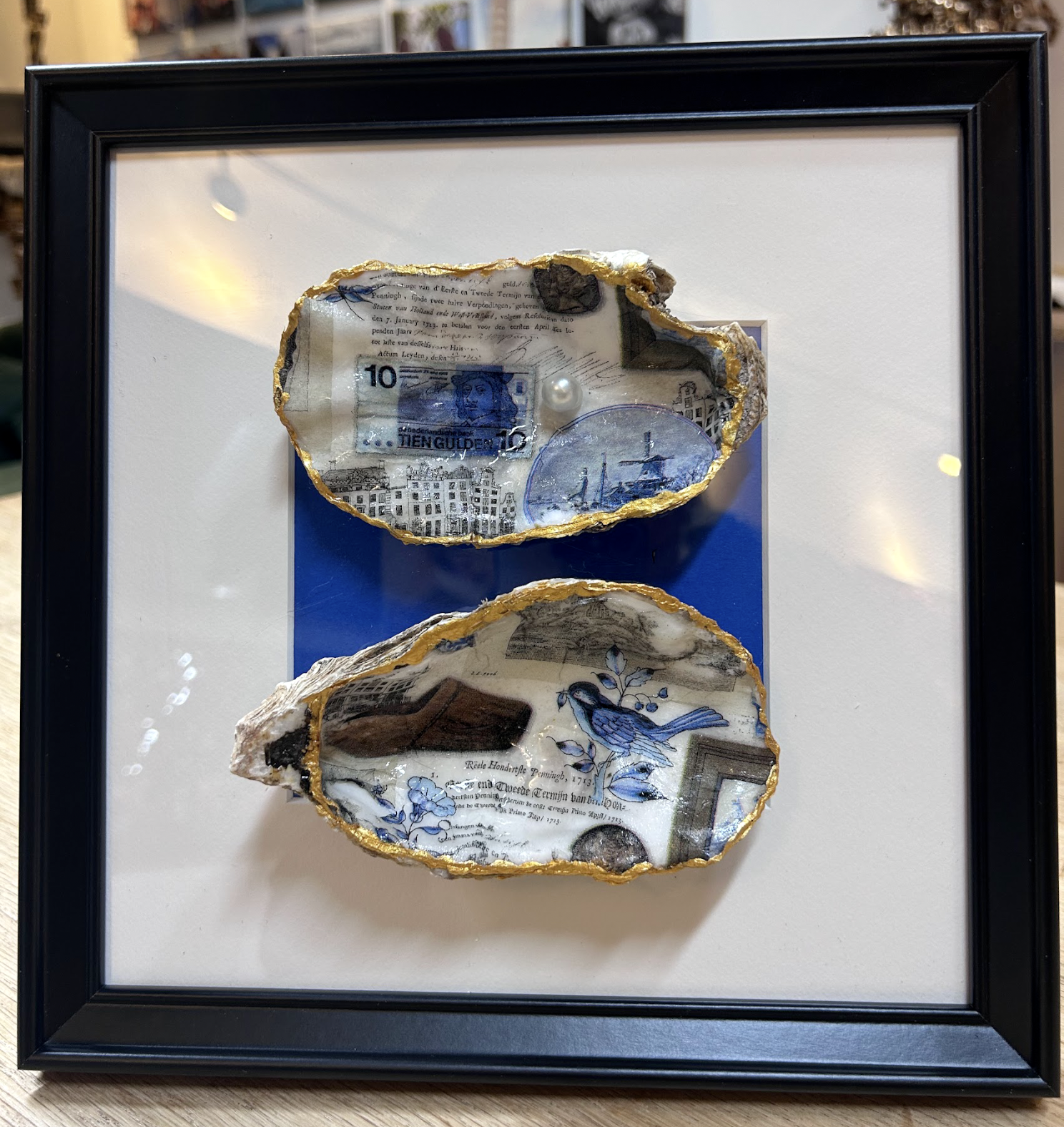 Ingelijst kunstwerk met twee schelpvormige keramische objecten op een witte passe-partout, tegen een blauwe achtergrond.