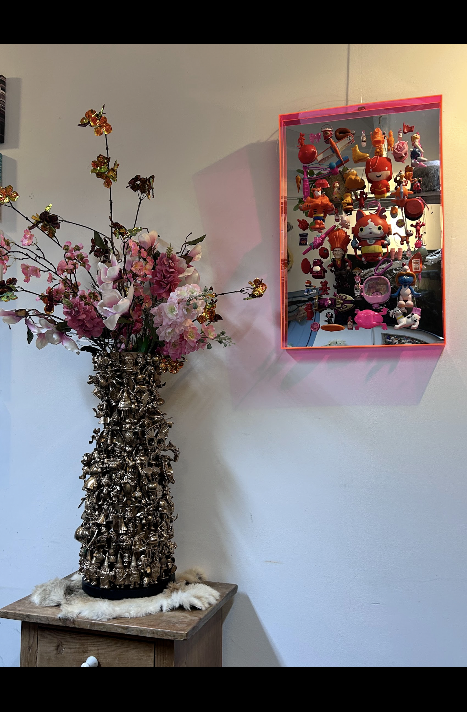 Een hoog bloemstuk op een tafel naast een roze ingelijste wanddecoratie met kleurrijke beeldjes.