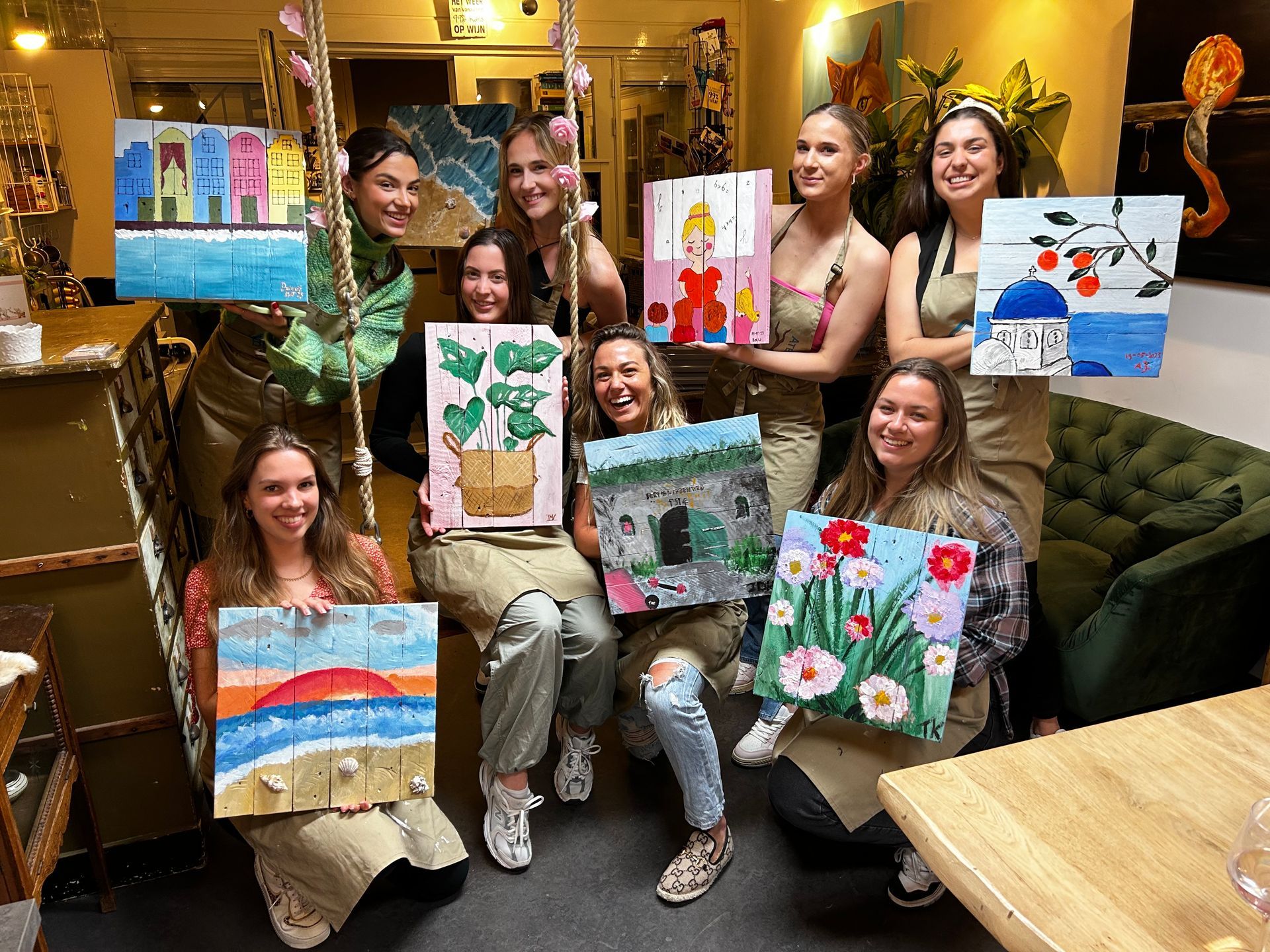 Een groep mensen poseert binnen met kleurrijke schilderijen, lachend en kunstwerken vasthoudend in een gezellige lounge.
