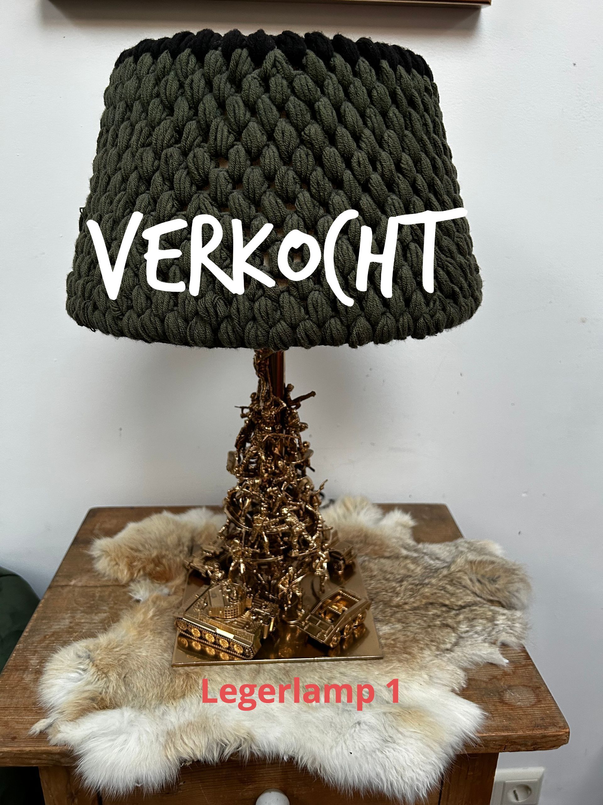 Een gebreide groene lampenkap op een gouden lampvoet met een pluizig crèmekleurig matje op een houten tafel.