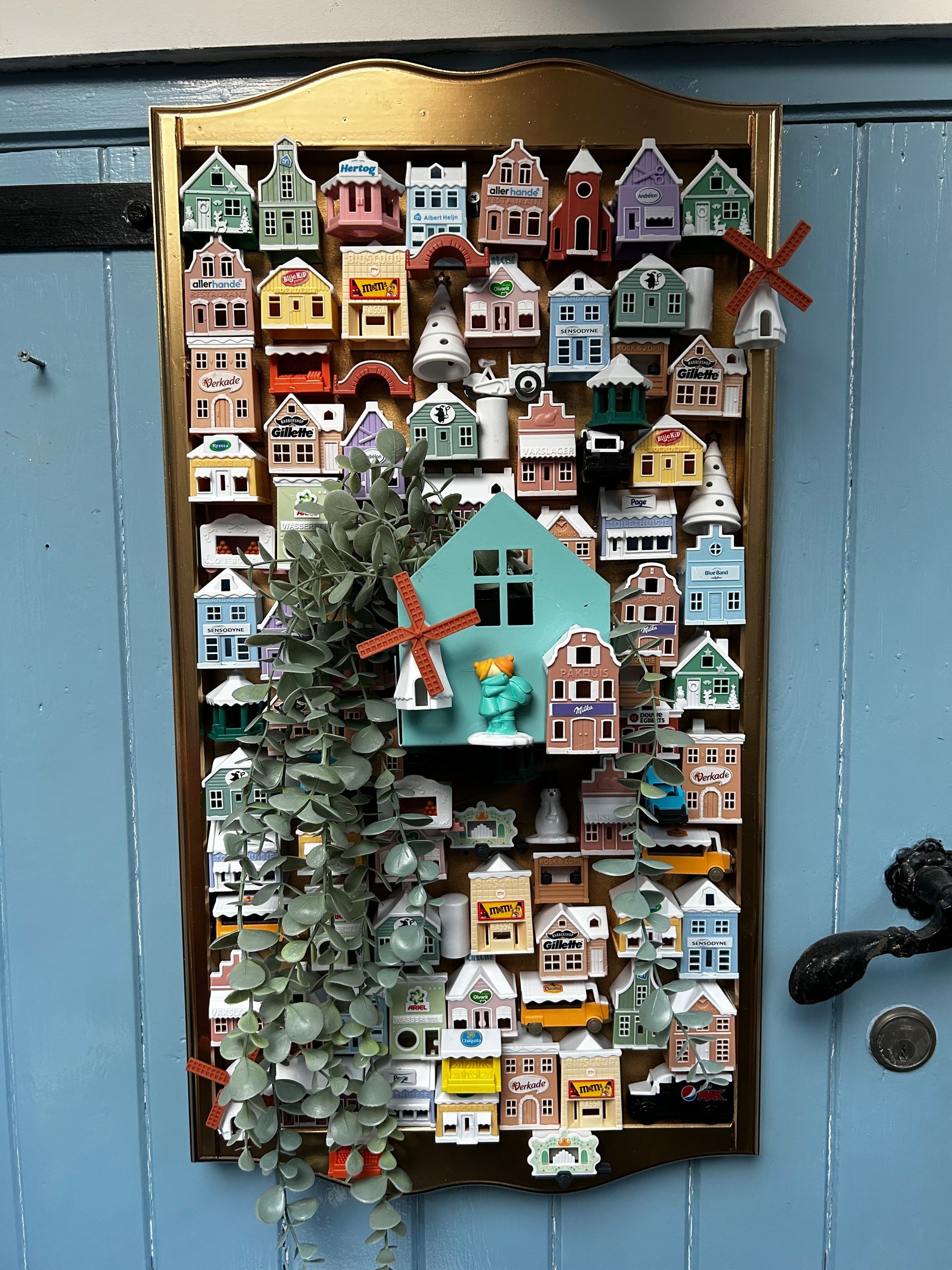 Kleurrijke miniatuurvogelhuisjes gerangschikt op een wandbord met kunstgroen en een turquoise middenstuk.