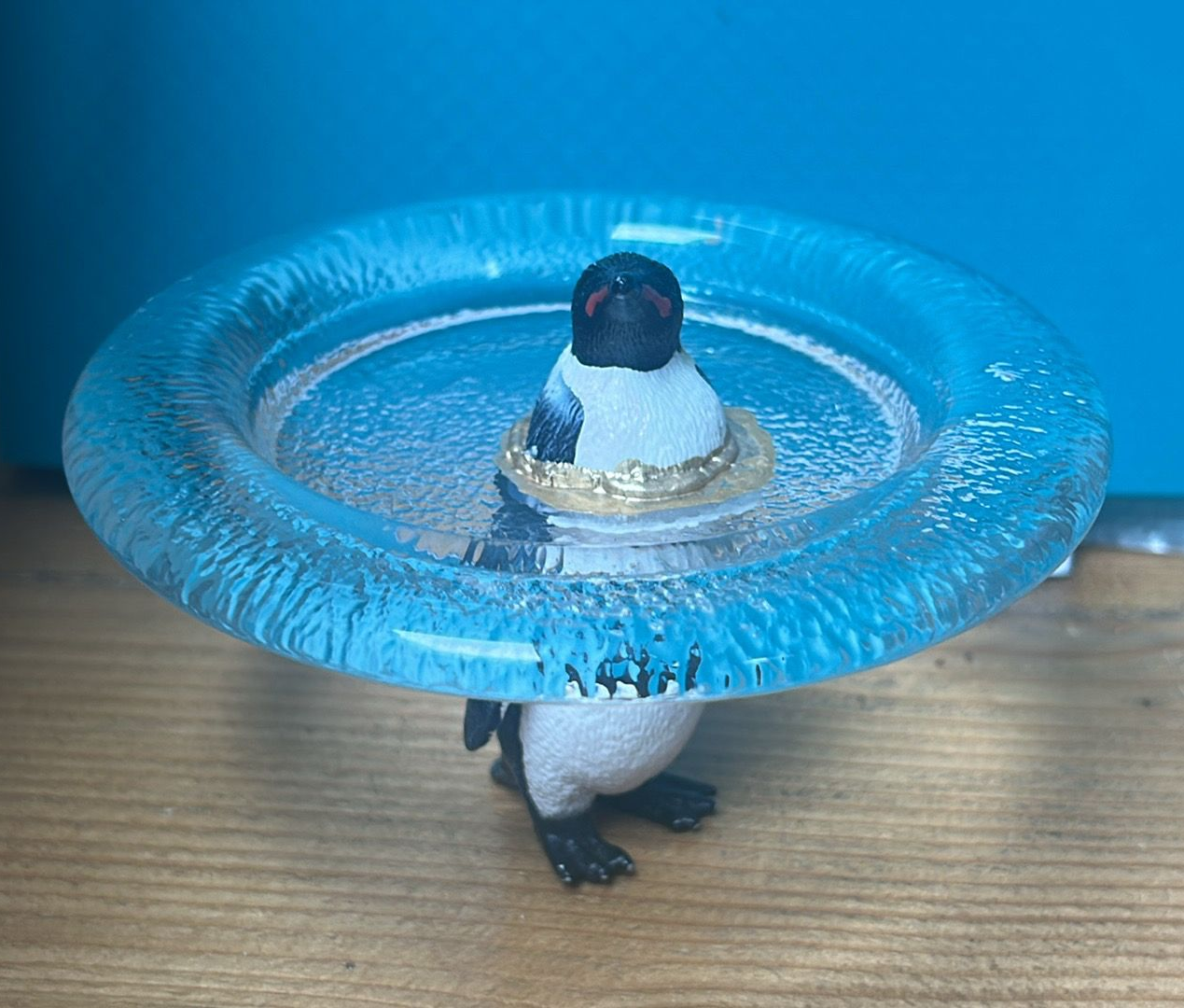 Speelgoedpinguïn met een blauwe plastic ring om zijn nek op een houten ondergrond.