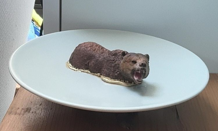 Een klein bruin pluche diertje rust op een wit bord op een tafel.