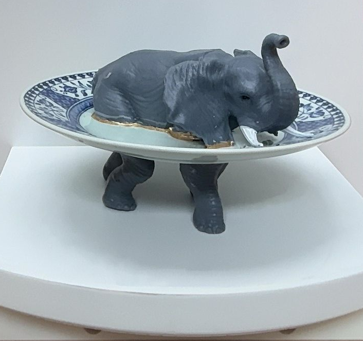 Een grijs olifantenbeeldje onder een decoratief bord, tentoongesteld op een witte draaibare standaard.