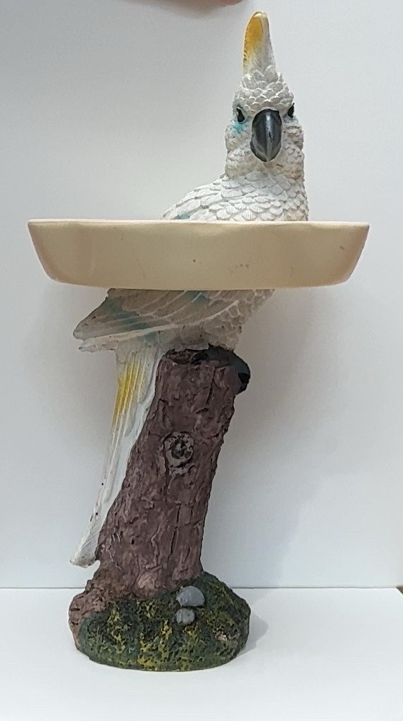 Beeldje van een witte kaketoe in een beige kom, geplaatst op een voetstuk van een boomstam met een mosachtige bodem.