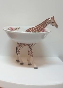 Een giraffenfiguurtje staat in een witte kom op een sokkel tegen een effen achtergrond.