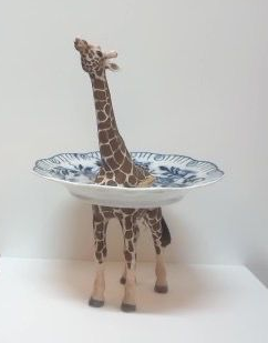 Een speelgoedgiraffe staat in een blauw-wit komvormig schaaltje, op een effen witte achtergrond.