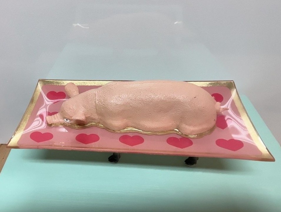 Roze varkenssculptuur op een dienblad, tentoongesteld in een museumachtige galerie tegen een lichtblauwe achtergrond