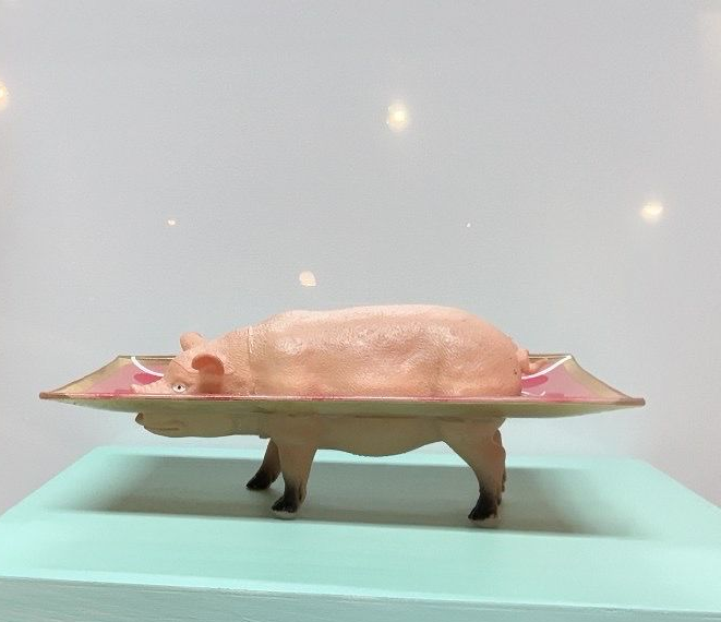 Roze varkenssculptuur op een dienblad, tentoongesteld in een museumachtige galerie tegen een lichtblauwe achtergrond