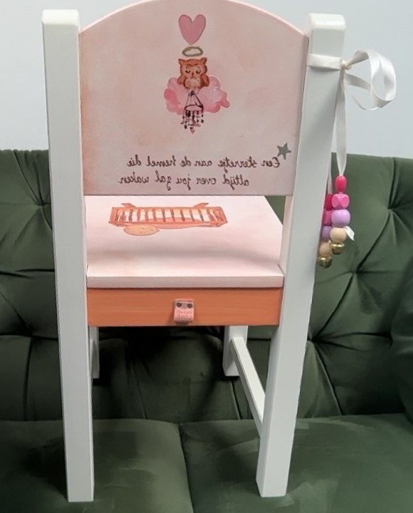 Roze kinderstoel met wit frame, afbeelding van een teddybeer en bungelende pomponkralen op een groene bank