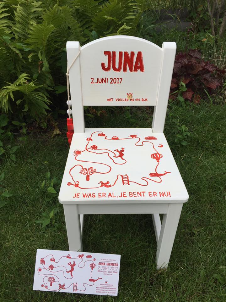 Witte stoel beschilderd met rode tekeningen en ‘JUNA’ op de rugleuning, opgesteld op gras met een bijpassend bordje.