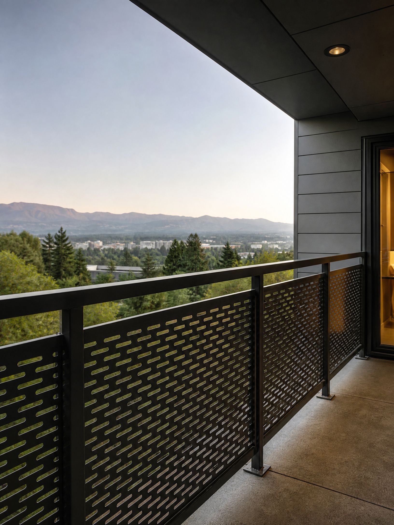 Balcony with black railing overlooking trees, hills, and a distant town at dusk