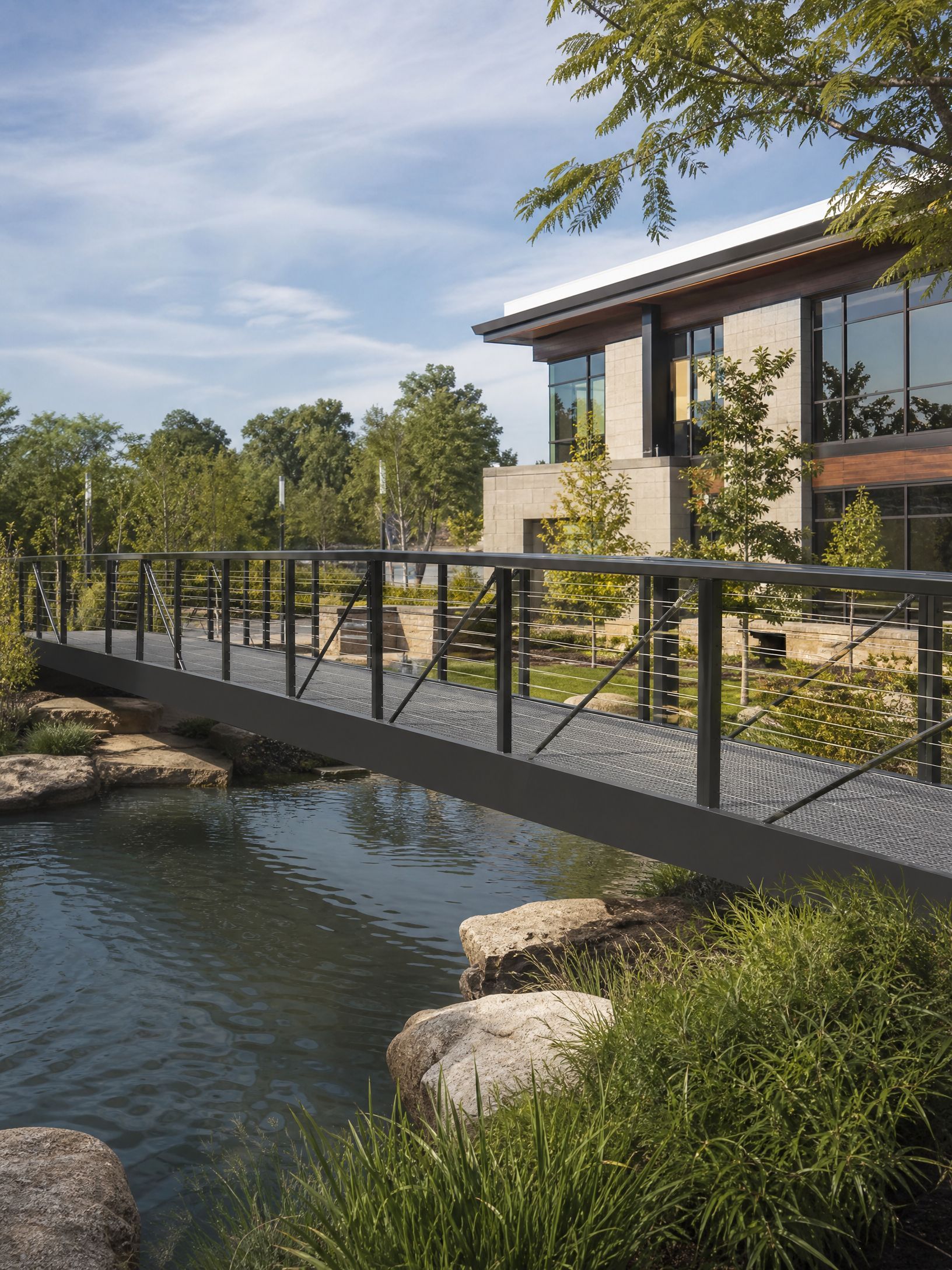 Modern house with a metal footbridge over a stream, surrounded by greenery and trees.