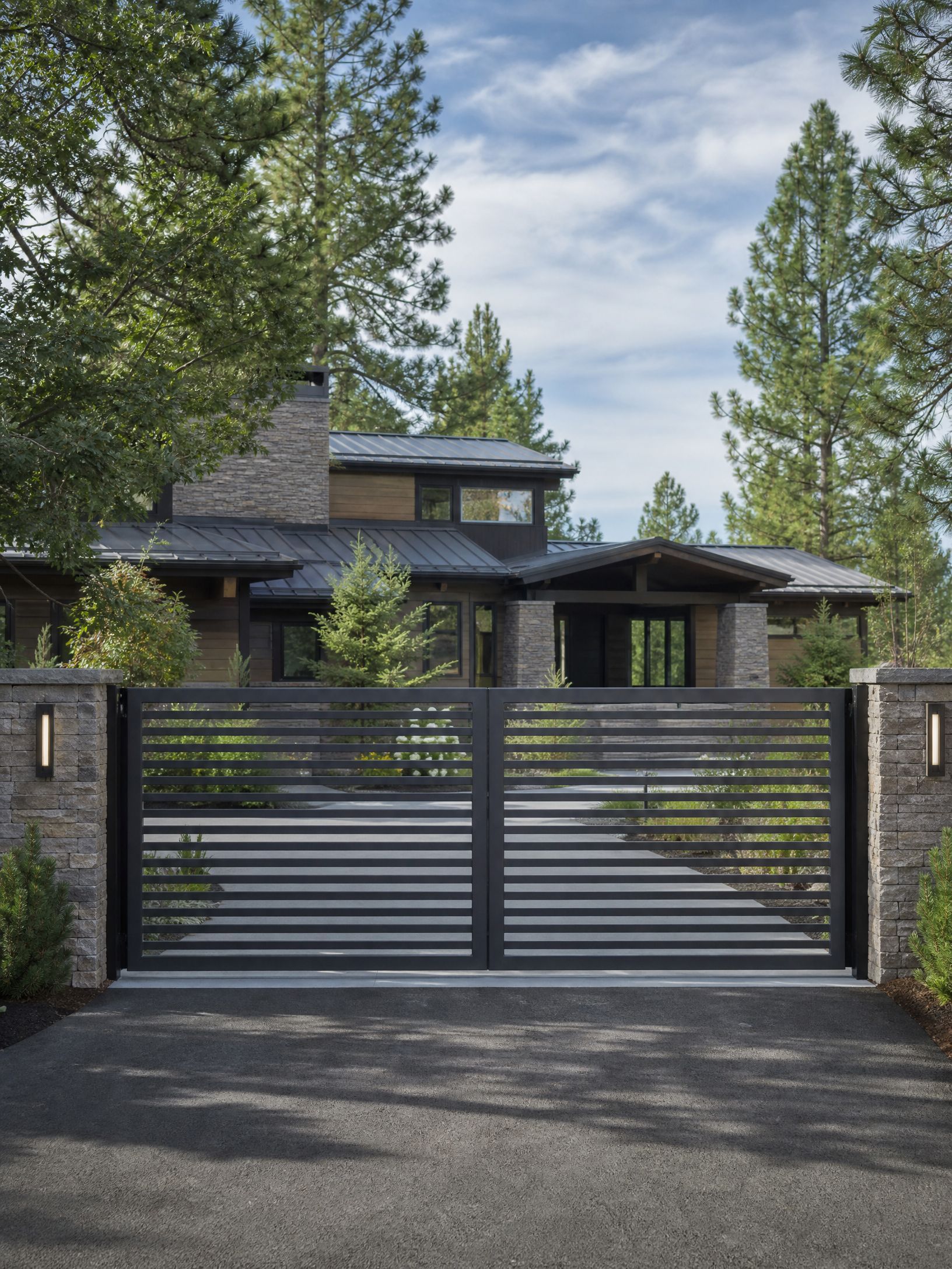 Modern house behind a gray metal gate, framed by tall pine trees.