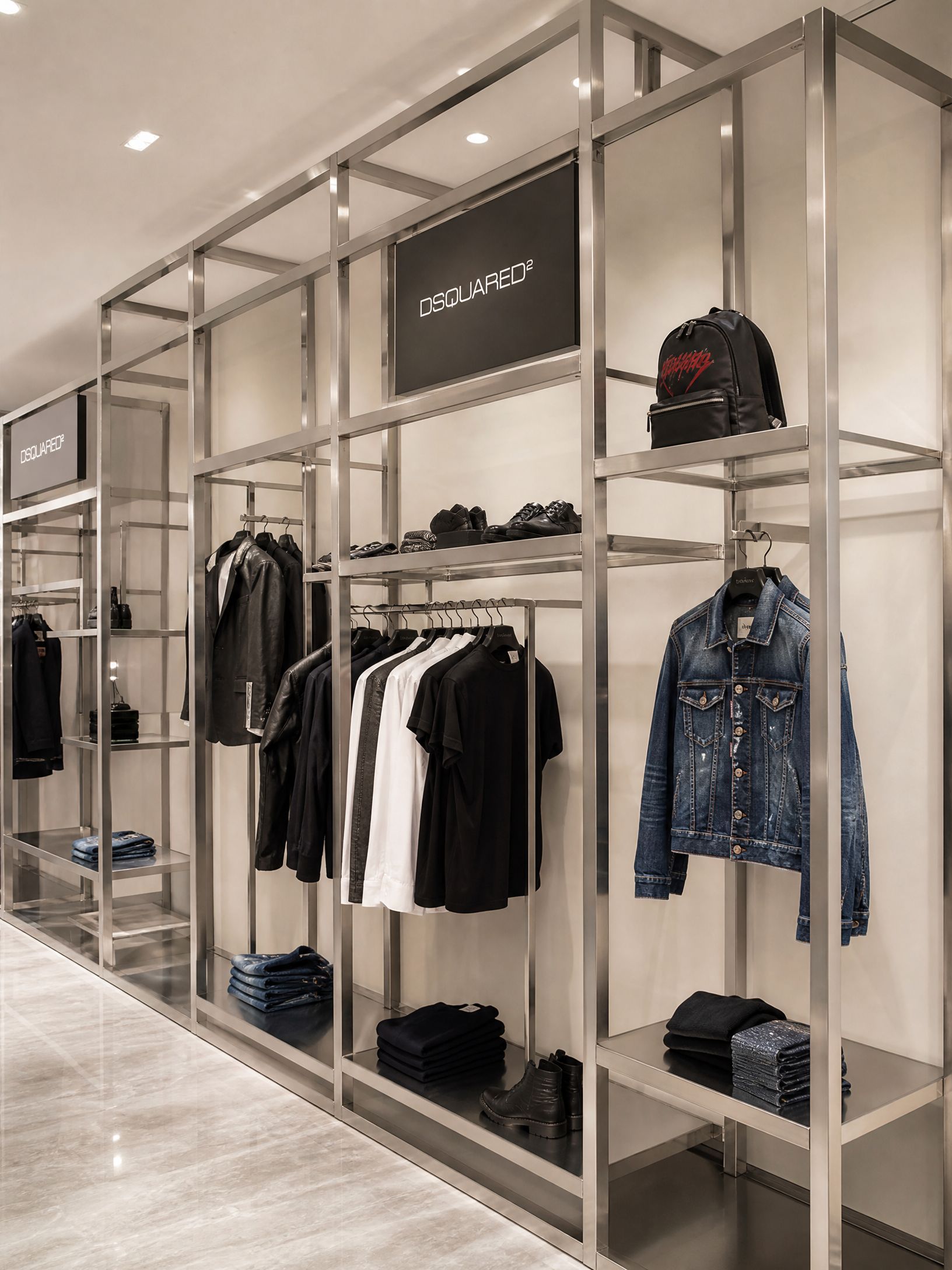 Modern clothing store display with metal racks, jackets, shirts, shoes, and handbags against a light wall