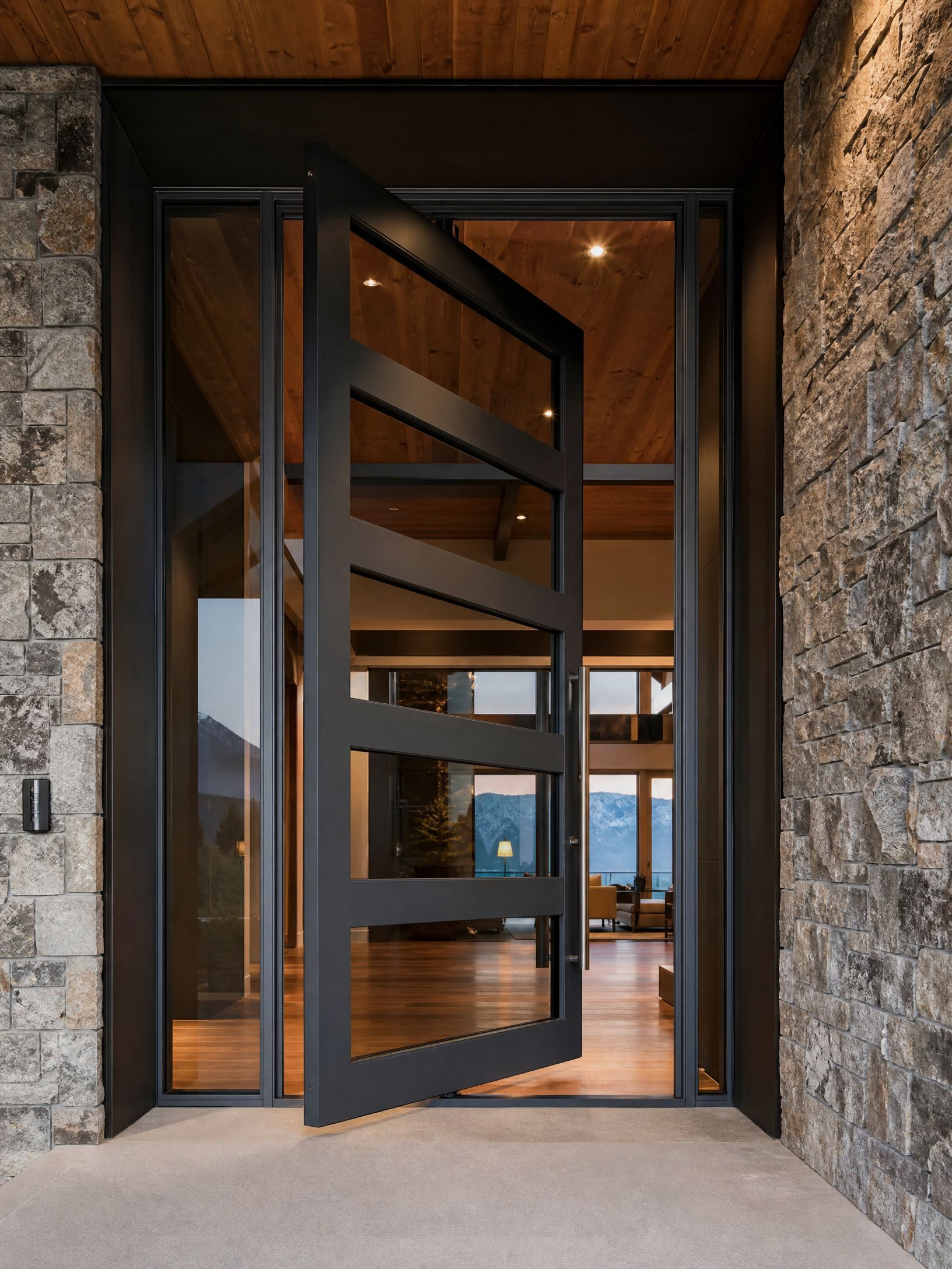 Modern black metal pivot door in a stone entryway with warm wood interior beyond