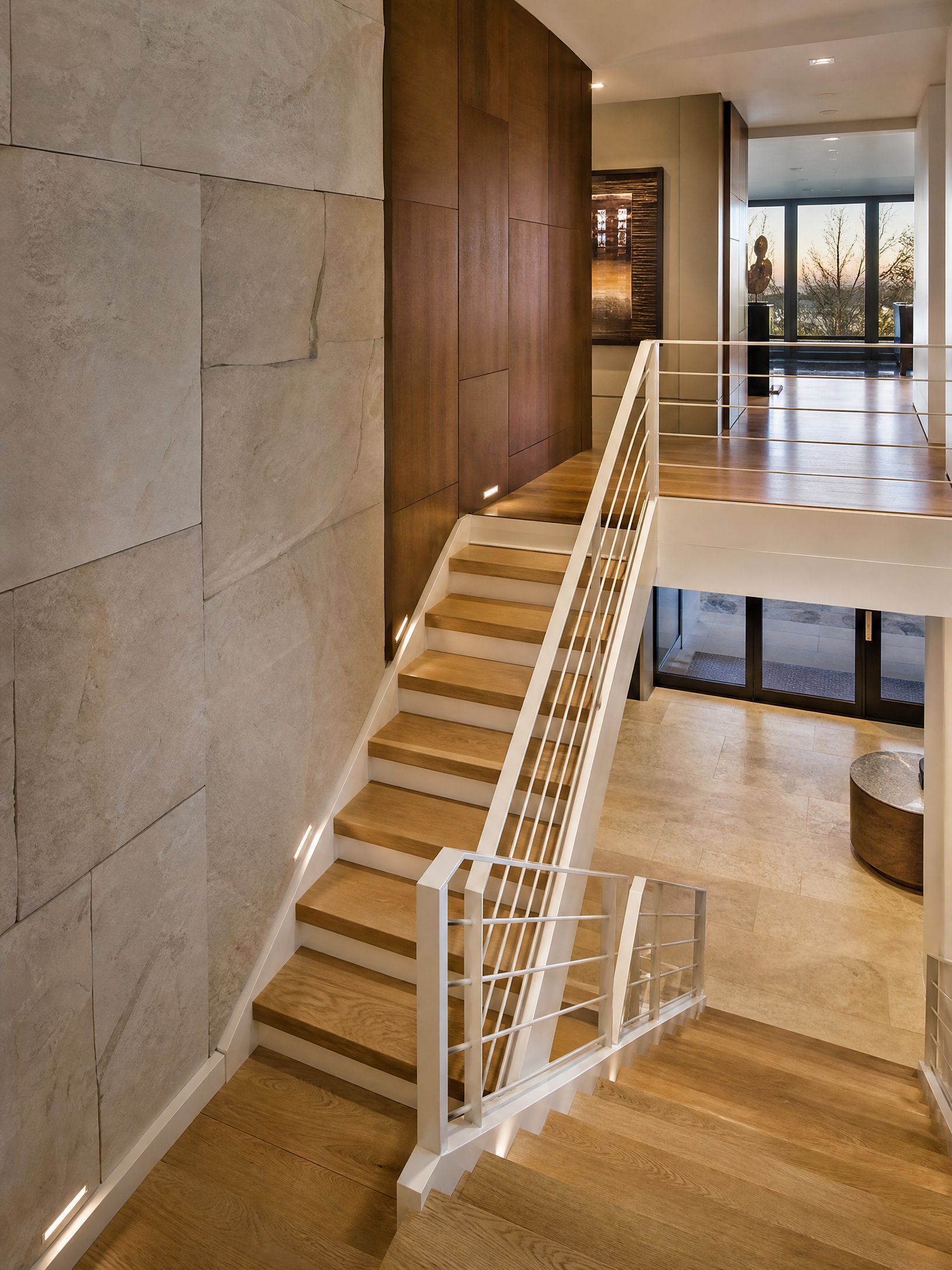 Modern lobby with a wooden staircase, glass railing, and stone walls.